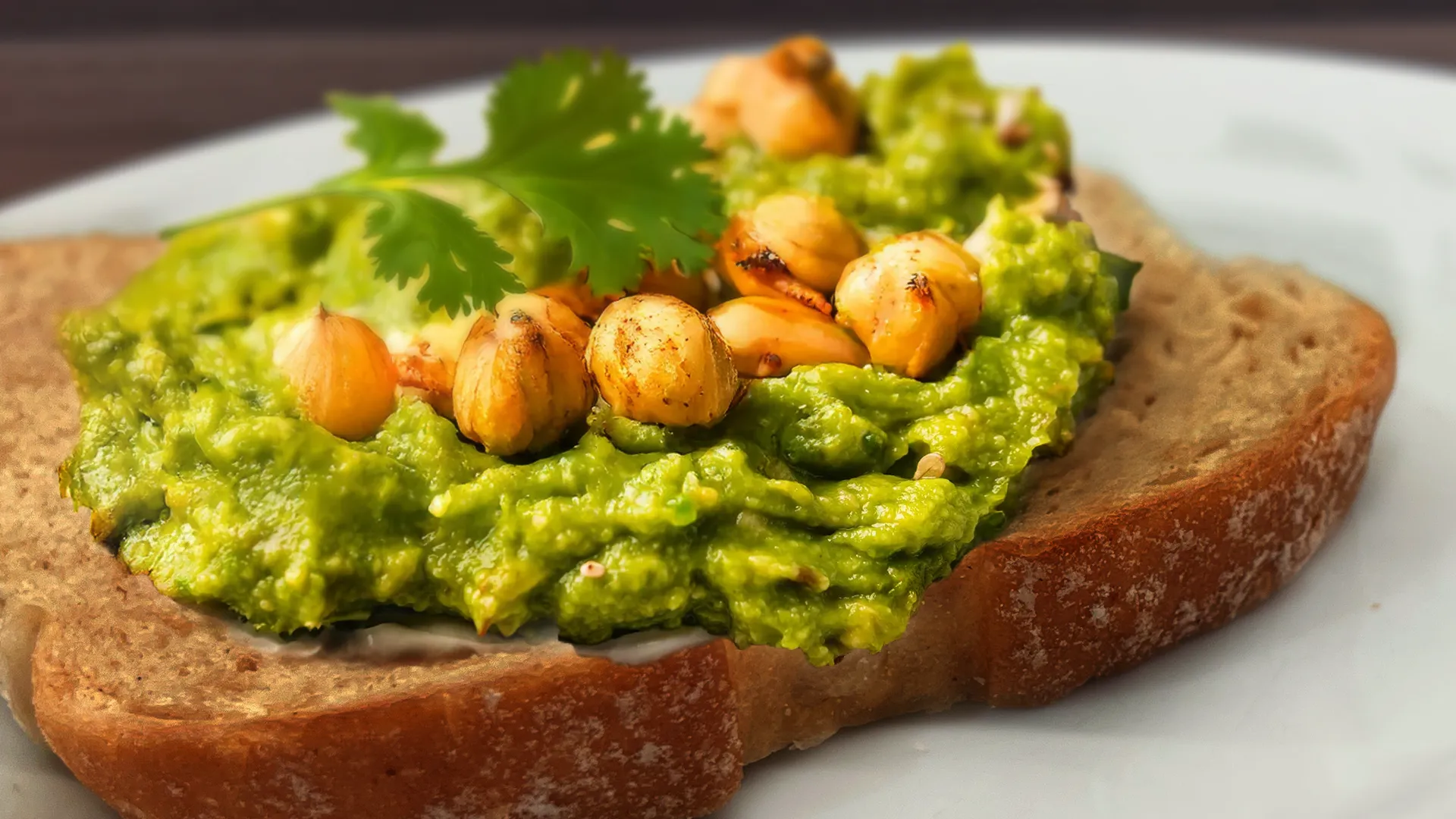 A close-up, eye-level shot of a slice of wholemeal toast on a white plate, generously topped with vibrant green smashed avocado and chickpeas. The chickpeas are lightly browned and seasoned, adding texture. A fresh sprig of cilantro garnishes the topping. The background is a blurred dark wood surface.