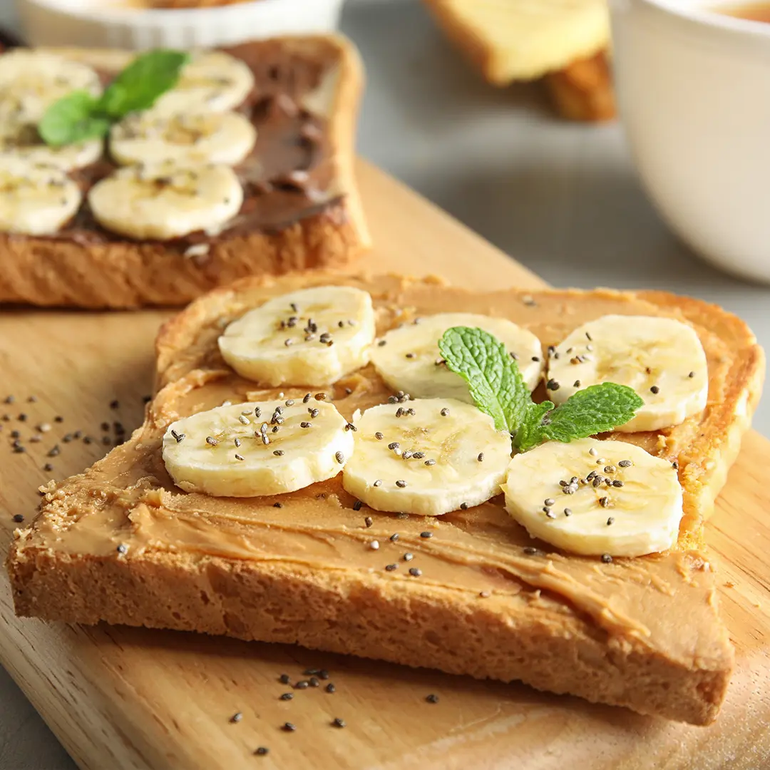 A top-down shot shows two slices of toast on a wooden cutting board. The front slice is topped with peanut butter, banana slices, and a sprinkle of chia seeds, garnished with a sprig of mint. The back slice has a chocolate spread, banana slices, and a sprig of mint. To the left, a small white bowl holds more chia seeds. In the blurred background, a white teacup with a spoon and a slice of plain toast are visible. The surface beneath the board is a light gray.