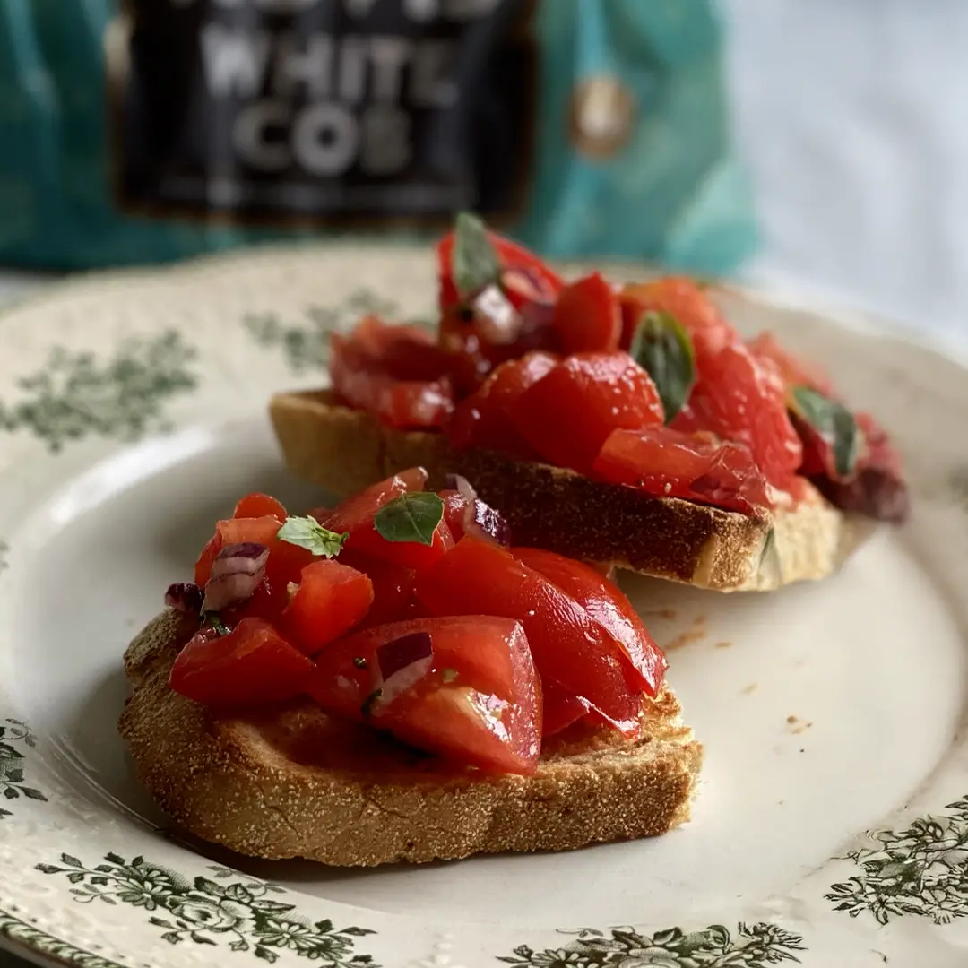 A close up of two half slices of Hovis White Cob on a floral plate, topped with a mix of diced tomatoes, onions and basil. In the background, a packaging of Hovis White Cob. 
