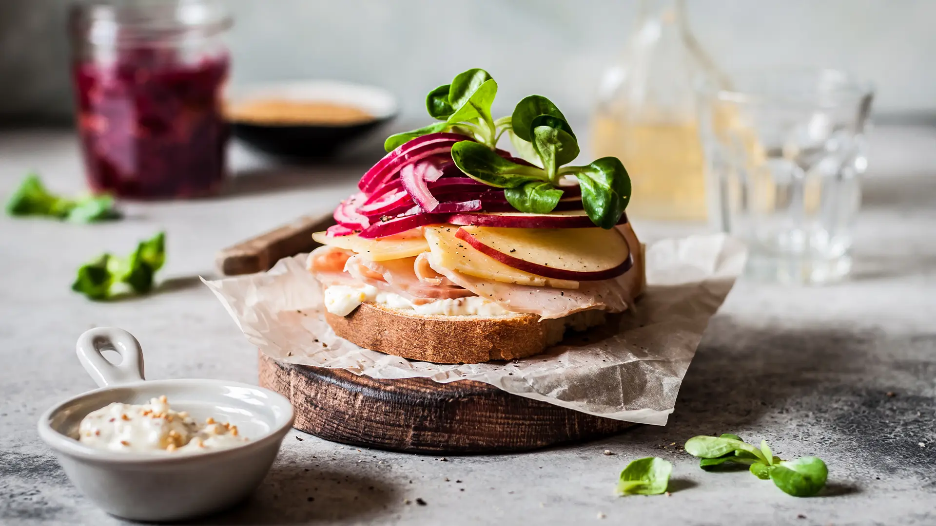 A delicious open-faced sandwich on a rustic wooden board, topped with sliced turkey or ham, cheese, thinly sliced red apple, and a vibrant pile of red onion and fresh greens. A small white bowl of sauce and a glass jar of what appears to be cranberry sauce are visible in the background, along with a bottle of oil or vinegar and a drinking glass. The setting appears to be a light and airy kitchen or dining area.