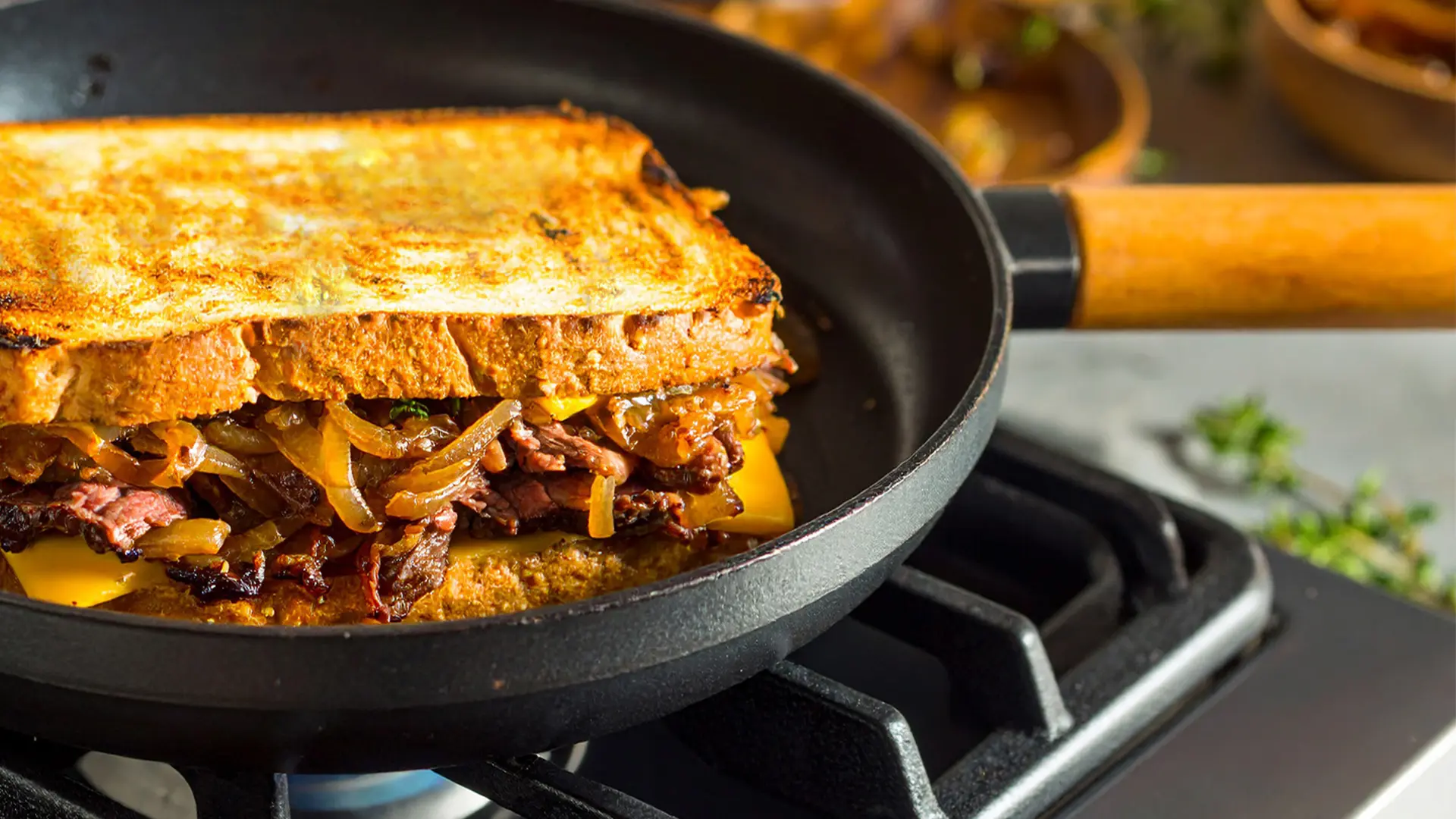 Beef & Caramelised Onion Grilled Cheese sandwich being toasted in a medium-sized frying pan, on a hob. In the background, a kitchen setting. 