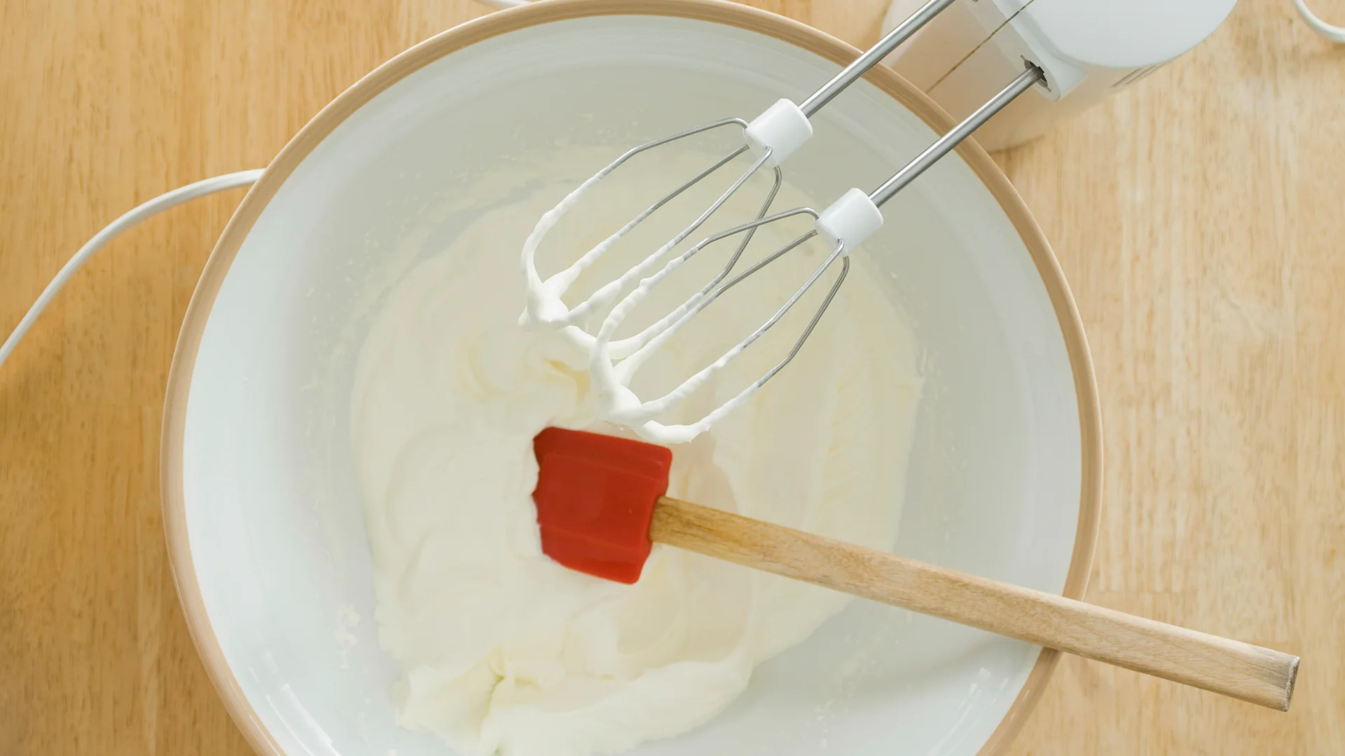 A top-down view of a medium bowl, filled with white fluffy cream on a wooden kitchen counter. Resting on the side, a red spatula and a mixer.