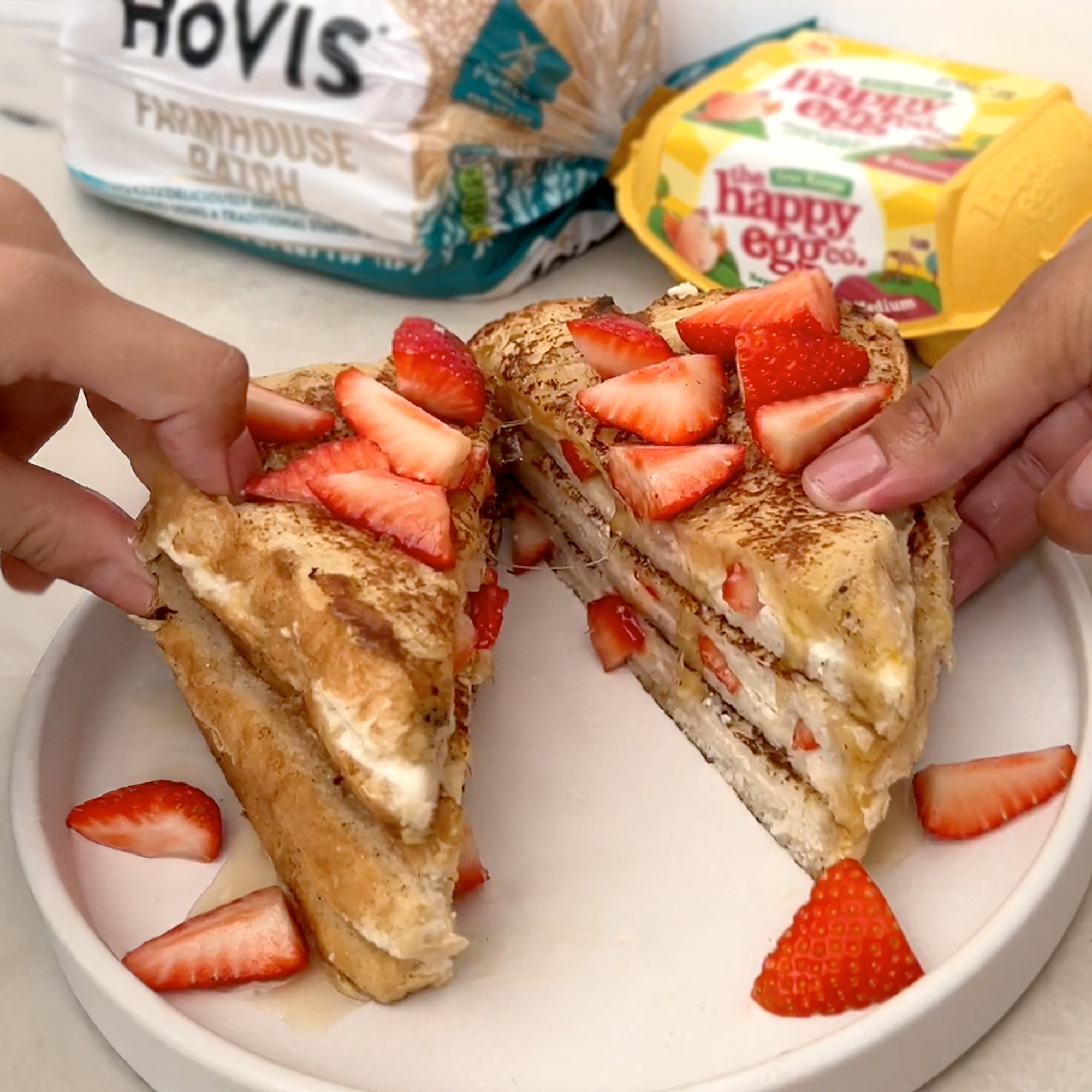 A white plate on a kitchen counter with 2 halves of a Strawberry Stuffed French Toast being pulled apart to reveal syrup and strawberries oozing out. A Hovis farmhouse batch loaf and a happy eggs pack can be seen in the background.