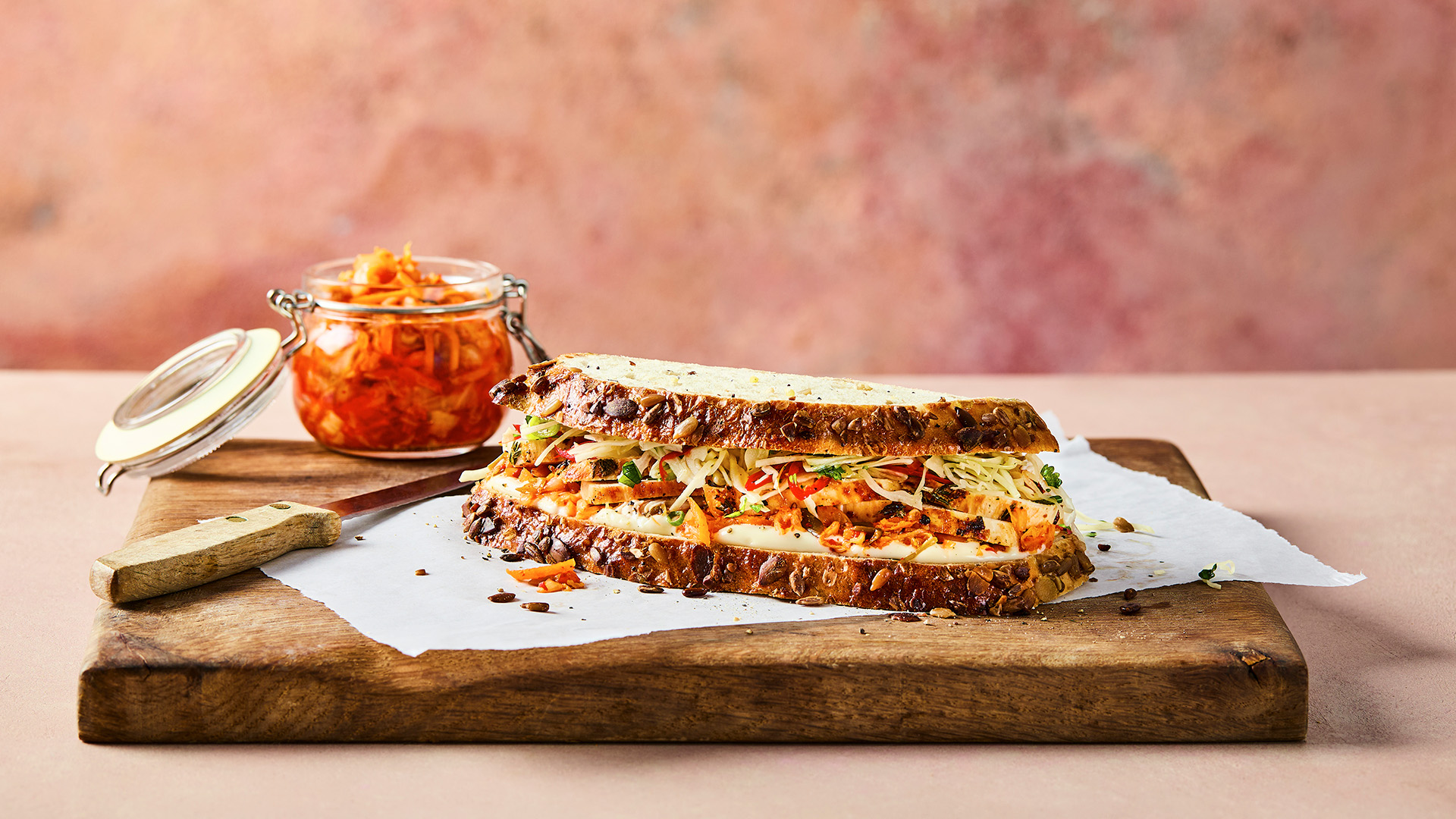 a seeded sourdough sandwich with slaw, kimchi and chicken breast pieces inside, sitting on a wooden chopping board with a bowl of leftover kimchi in the background 