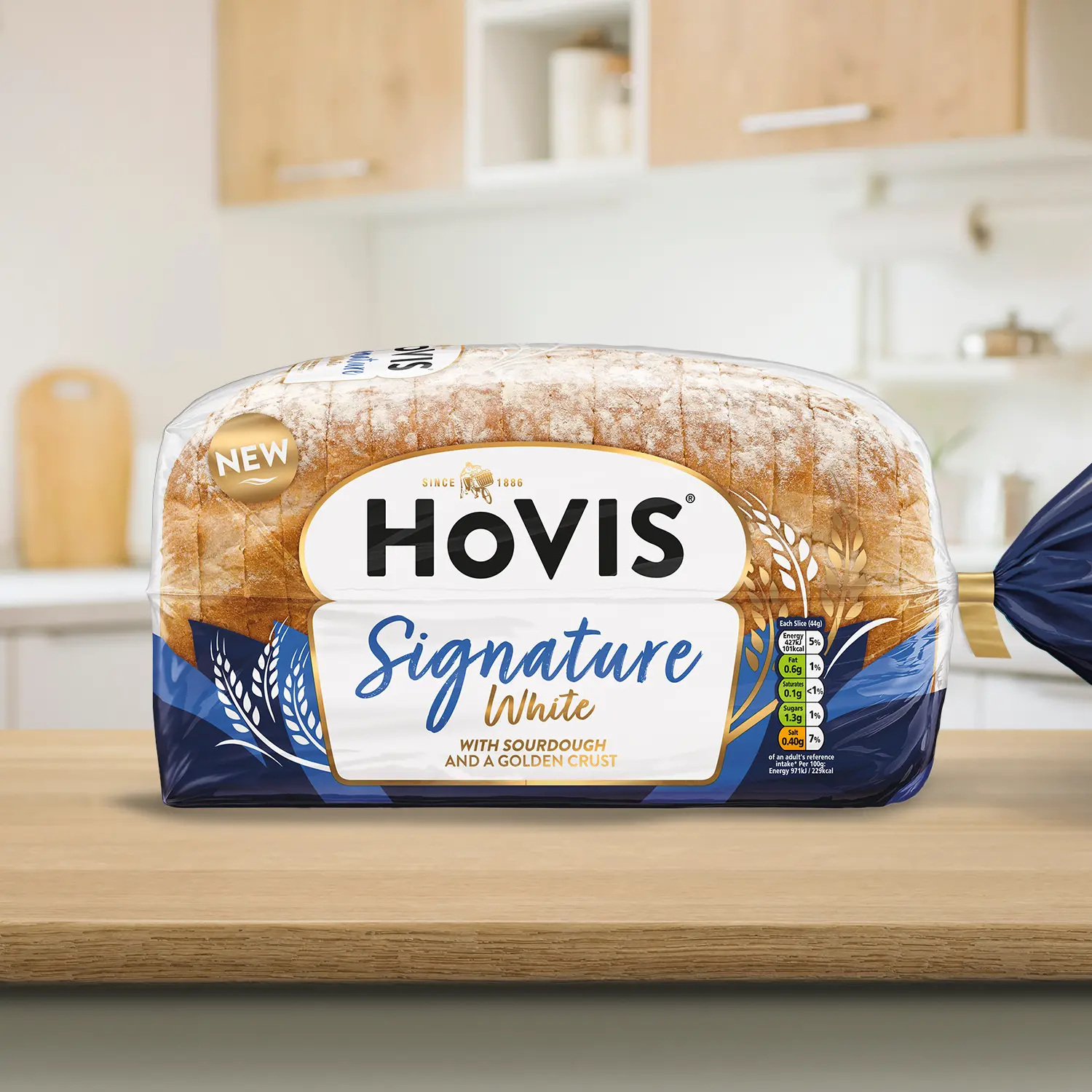 A loaf of Hovis Signature White bread with sourdough and a crust dusted with flour, displayed on a wooden kitchen countertop. The packaging features a white and navy design with a gold trim and nutrition information. A 'NEW' label is shown on the top corner of the pack