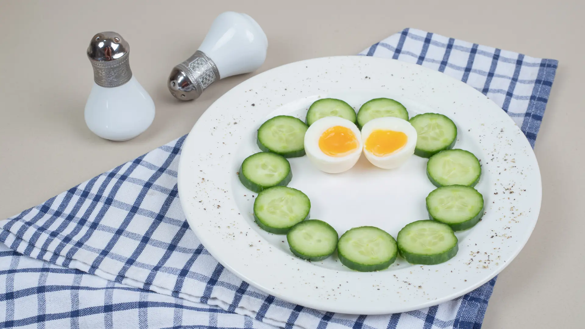 A soft boiled egg cut in half surrounded by a circle of cucumber slices on a plate, with unique salt and pepper shakers.