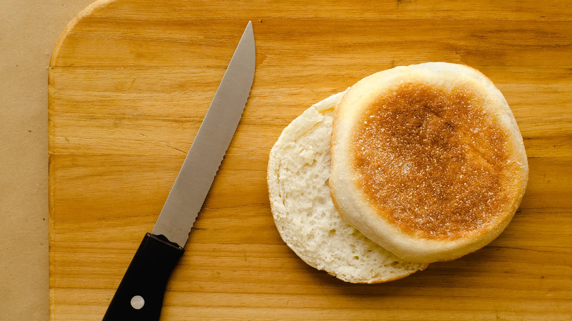 A delicious Hovis English Muffin sliced in half on top of a wooden chopping board, with a sharp knife on the left hand side. 
