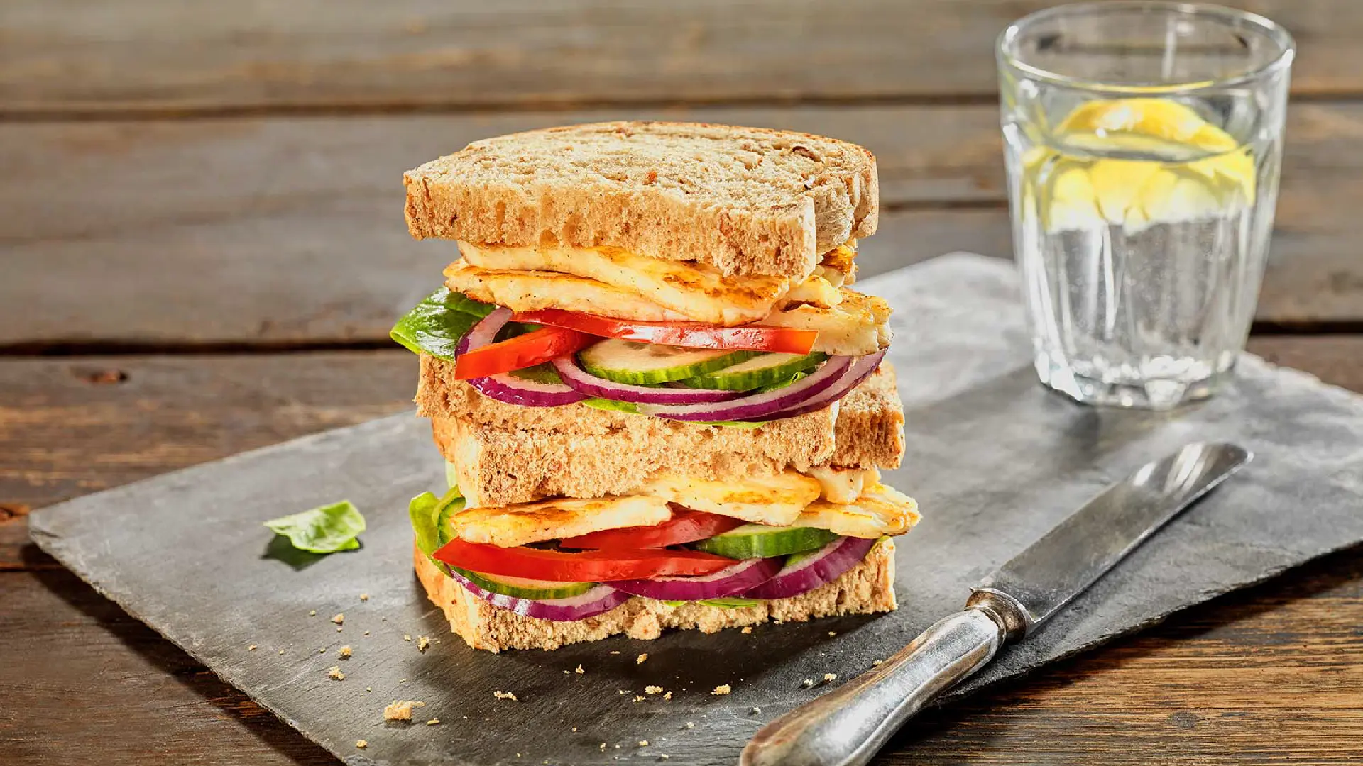 Two Hovis Granary 400g sandwiches stacked, filled with slices of red onion, cucumber, red pepper, and halloumi between brown bread slices.