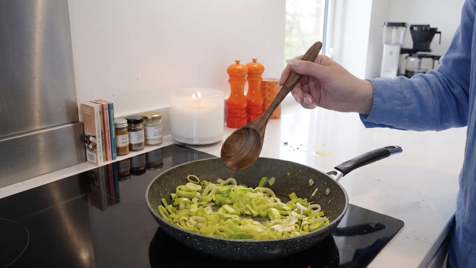 A person holds a wooden spoon over a frying pan filled with chopped green leeks cooking on a modern kitchen stovetop.
