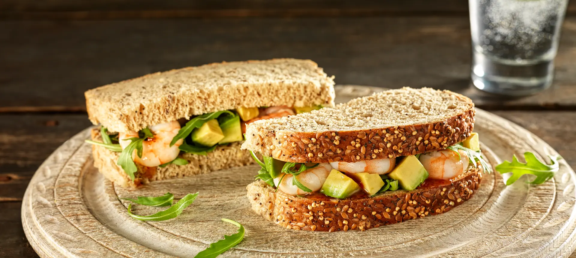 A sandwich made with Hovis Seeded Batch bread, sliced in half and filled with diced avocado, prawns, and rocket leaves, on a beige plate.