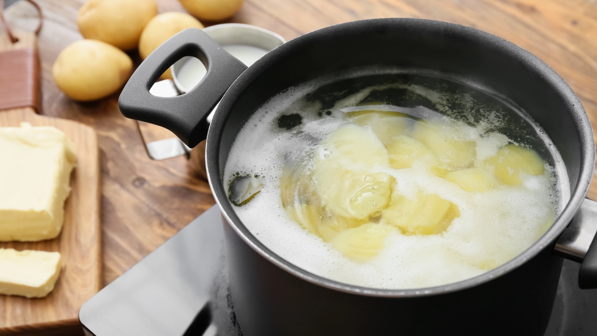A large pot filled with simmering water and sliced potatoes cut lengthwise rests on the hob. In the background are small potatoes and a block of butter.
