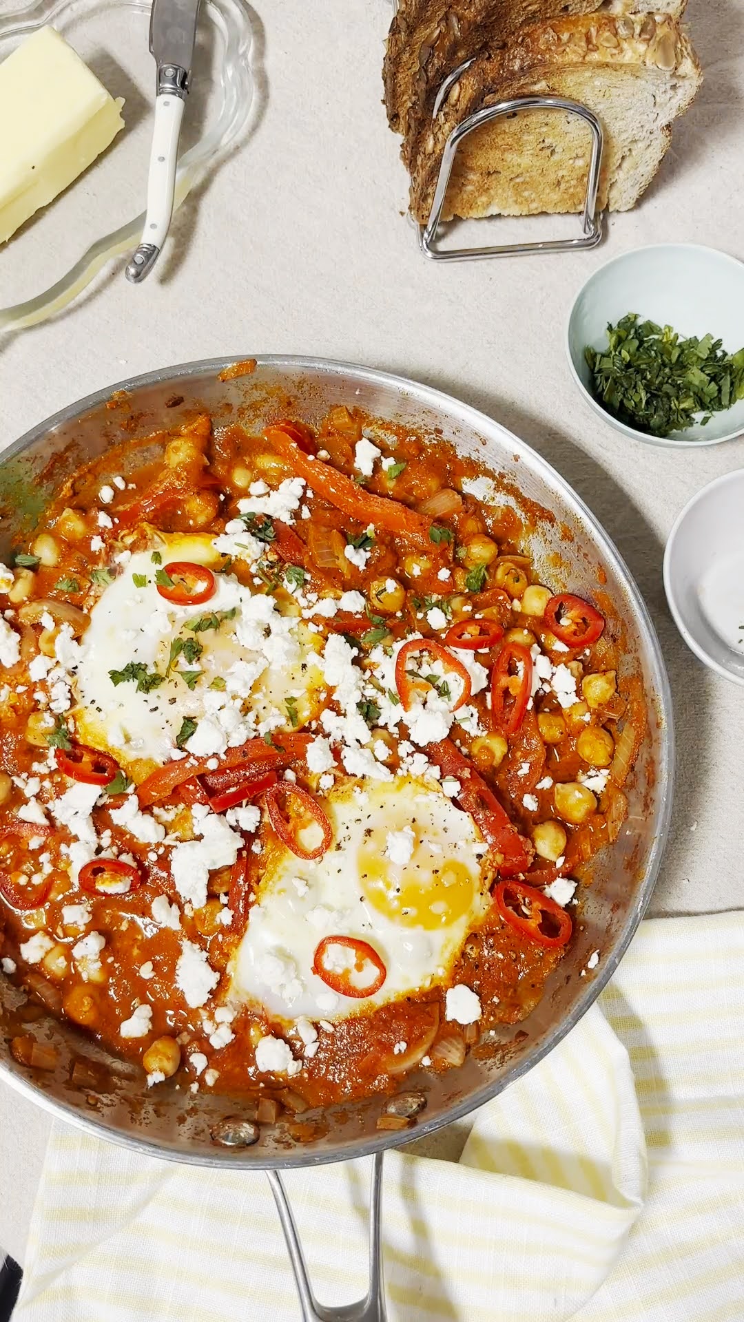 A skillet filled with vibrant shakshuka sits on a light countertop, featuring poached eggs nestled in a rich tomato sauce with chickpeas, red peppers, onions, and spices. The dish is topped with crumbled feta, sliced red chillies, and fresh herbs for extra colour and flavour. Nearby, a rack of toasted seeded bread, a dish of butter, and a small bowl of chopped herbs complete the cosy breakfast setup, ready to be served.