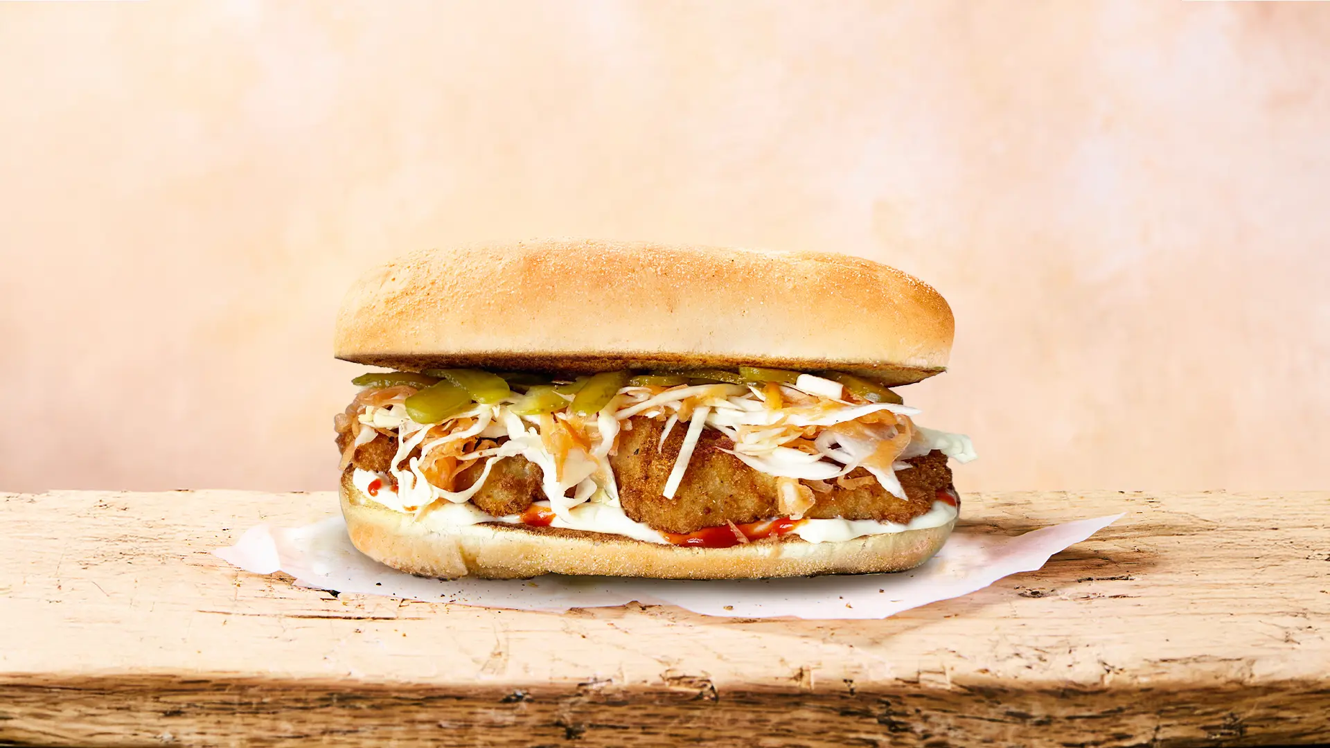 A fresh Hovis Buttermilk Sub Rolls sandwich, filled with pieces of fried chicken, grated white cabbage and carrots, thin slices of gherkin, and sauce rest on top of a napkin in a light beige background. 