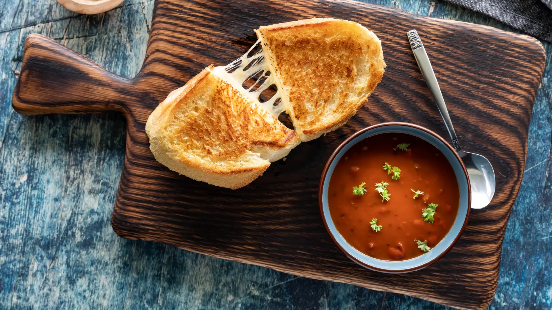 A top-down view of a bowl filled with tomato soup garnished with green herbs, alongside a sliced cheese toastie made with Hovis White Half Cob.