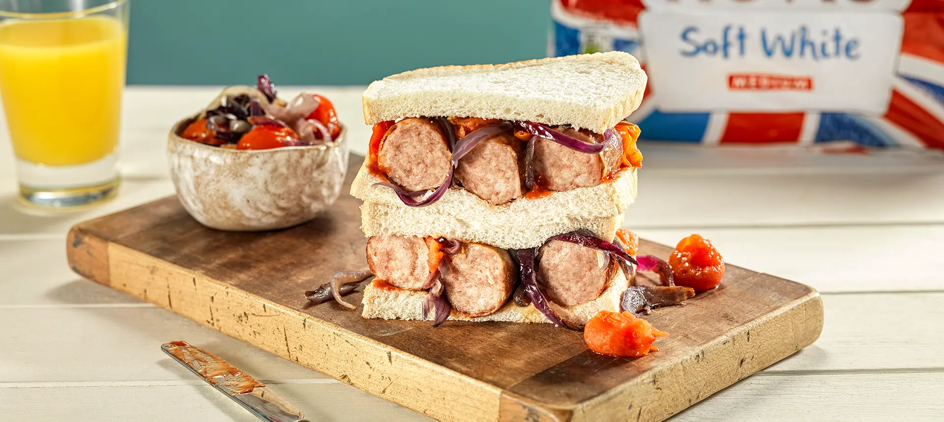 Two halves of a sandwich made with Hovis Soft White Medium Sliced bread, stacked and filled with red onions, sausage, and roasted tomatoes, on a wooden chopping board.
