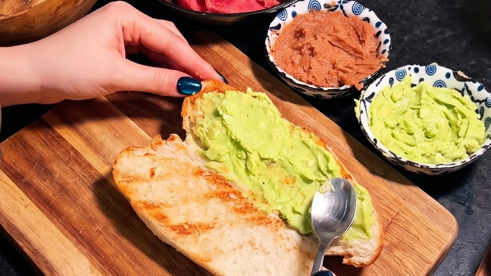 A spoon spreads mashed avocado onto toasted bread, next to bowls of green and red spreads on a wooden cutting board.