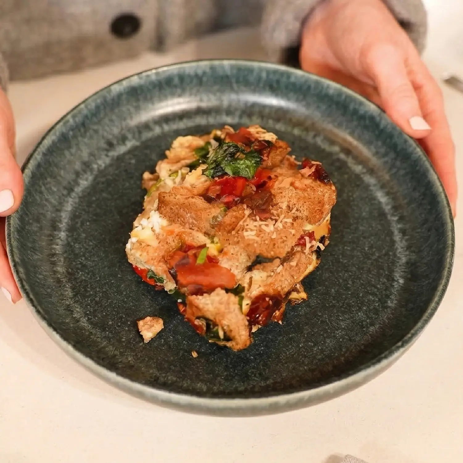A slice of savory breakfast strata with crusty bread, spinach, and red peppers served on a dark textured ceramic plate.