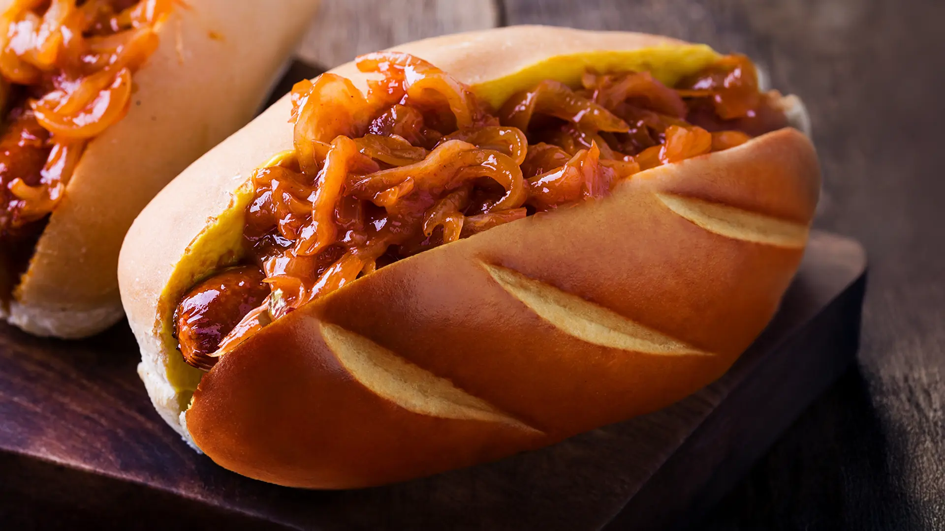 A delicious hot dog in a lightly toasted bun, generously topped with an abundance of glistening caramelized onions. The bun has decorative diagonal slashes on its golden-brown crust. A hint of another hot dog is visible in the blurred background on the left. The hot dog and bun are resting on a rustic wooden board, with a dark, blurred background.