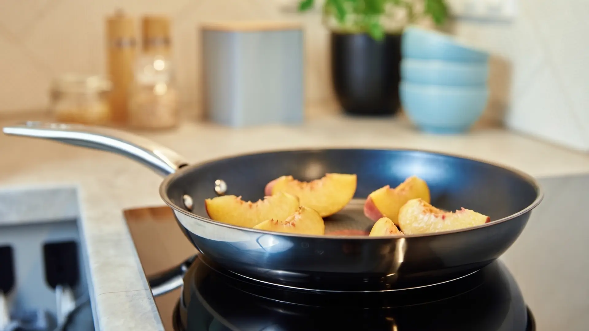 Slices of yellow peach cooking in a black frying pan on a stovetop burner, with a blurred kitchen background.
