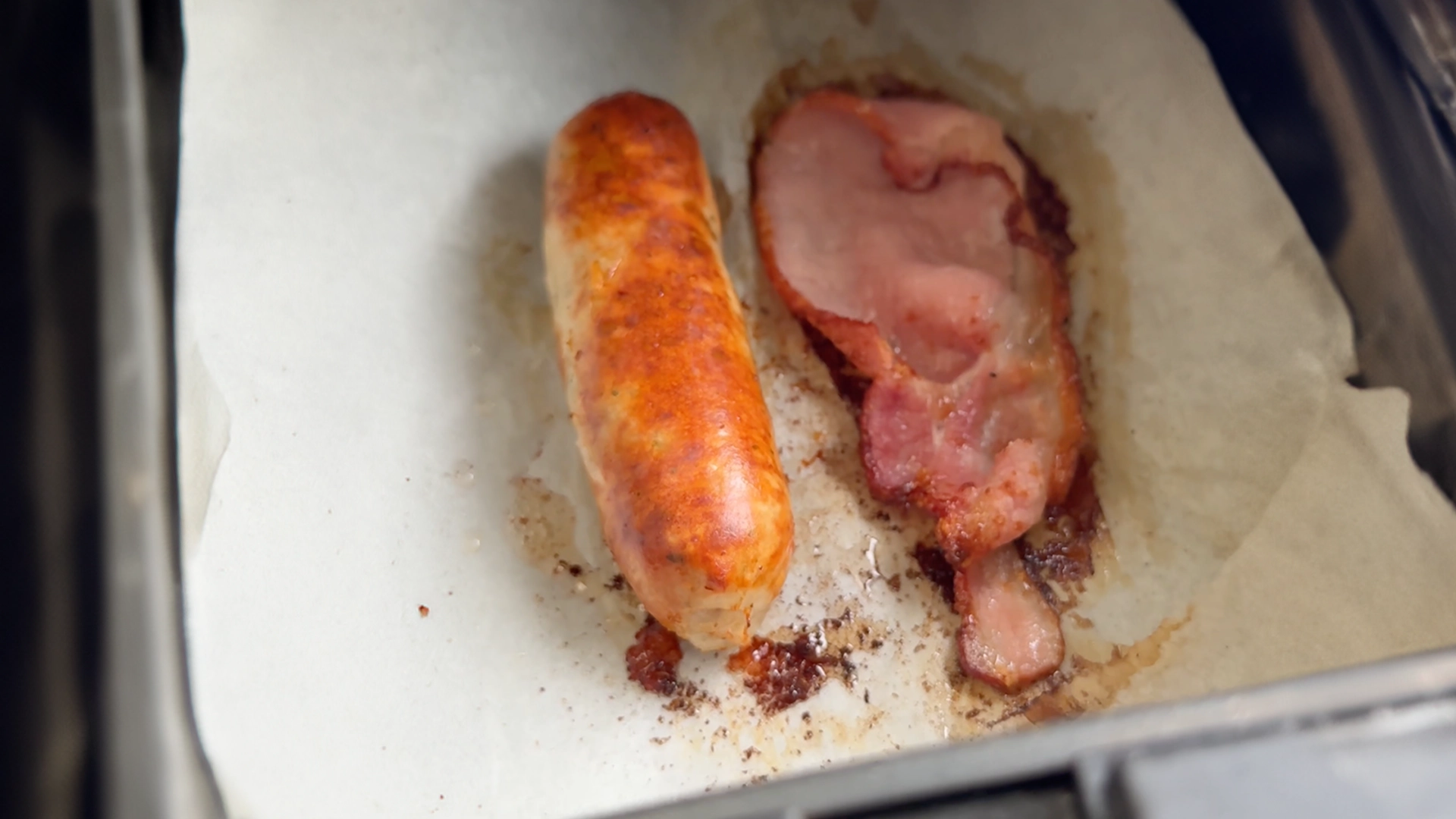 One sausage and a bacon rasher, golden-brown, on baking paper in a metal tray, being taken out of the air fryer or oven.