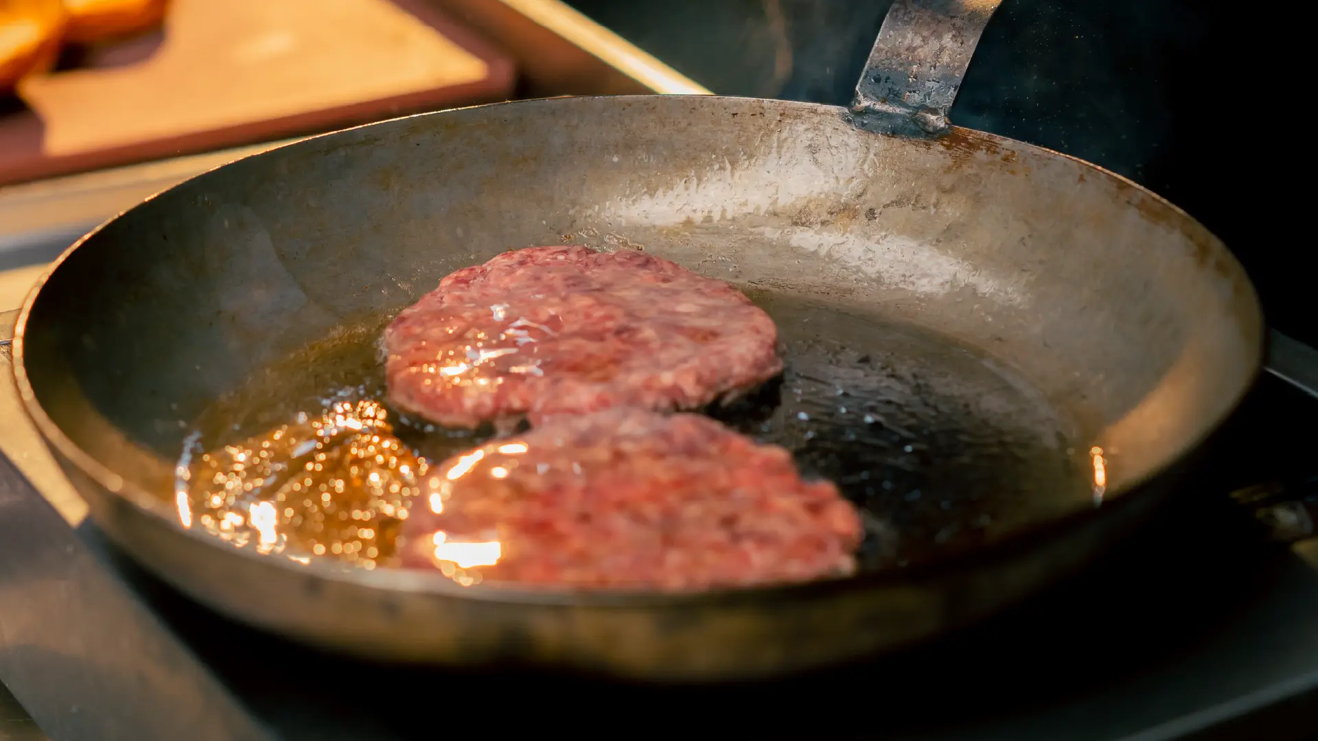 An angles view of a medium sized frying pan on top of a kitchen hob, with two meat patties being cooked with some oil.