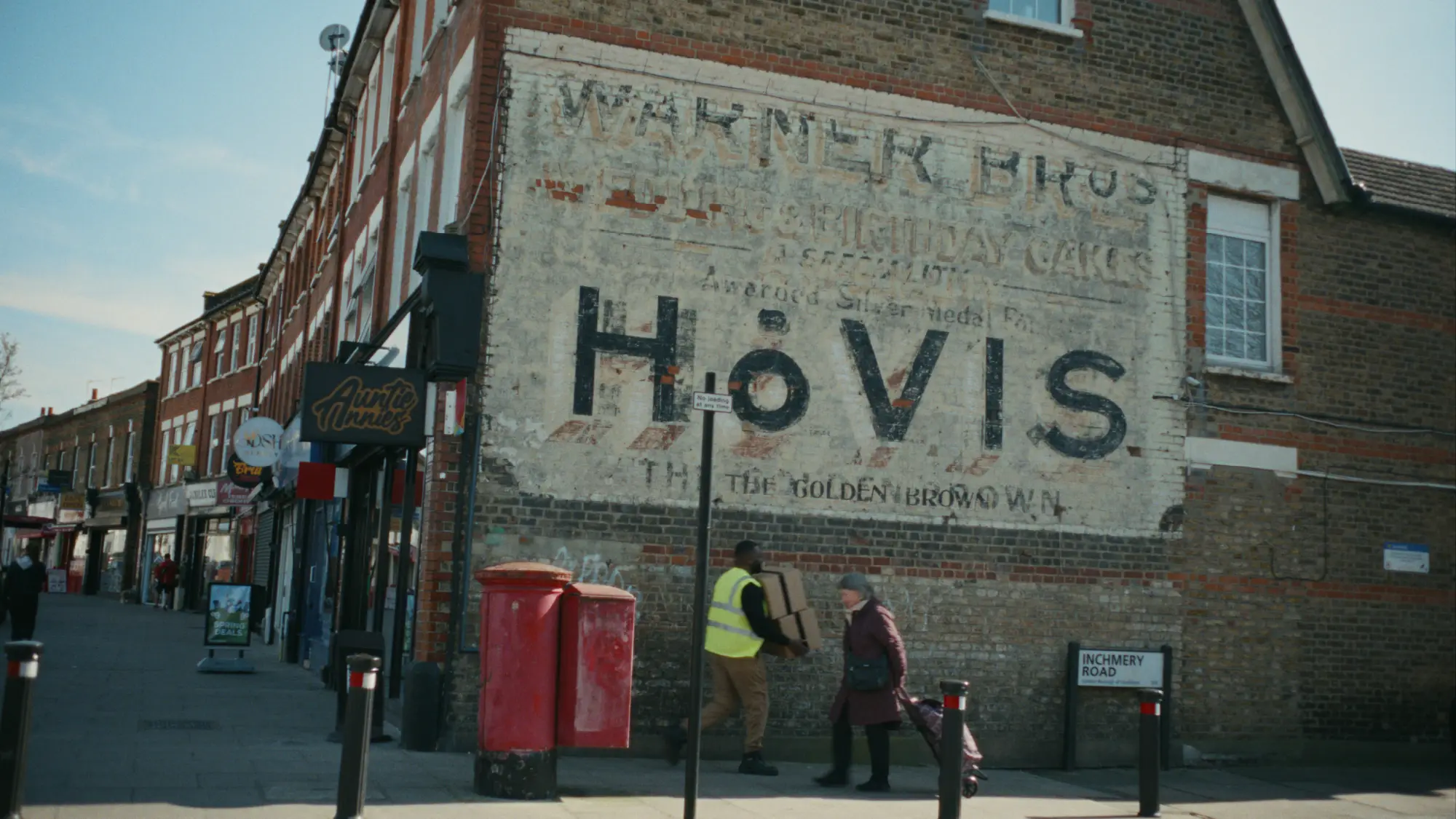 Vintage Hovis ghost sign on a brick wall in a British high street, featuring the iconic bread brand’s faded advertisement. Local shops, pedestrians, and a red postbox appear in the foreground on Inchmery Road, capturing everyday life and Hovis’s heritage in UK communities.
