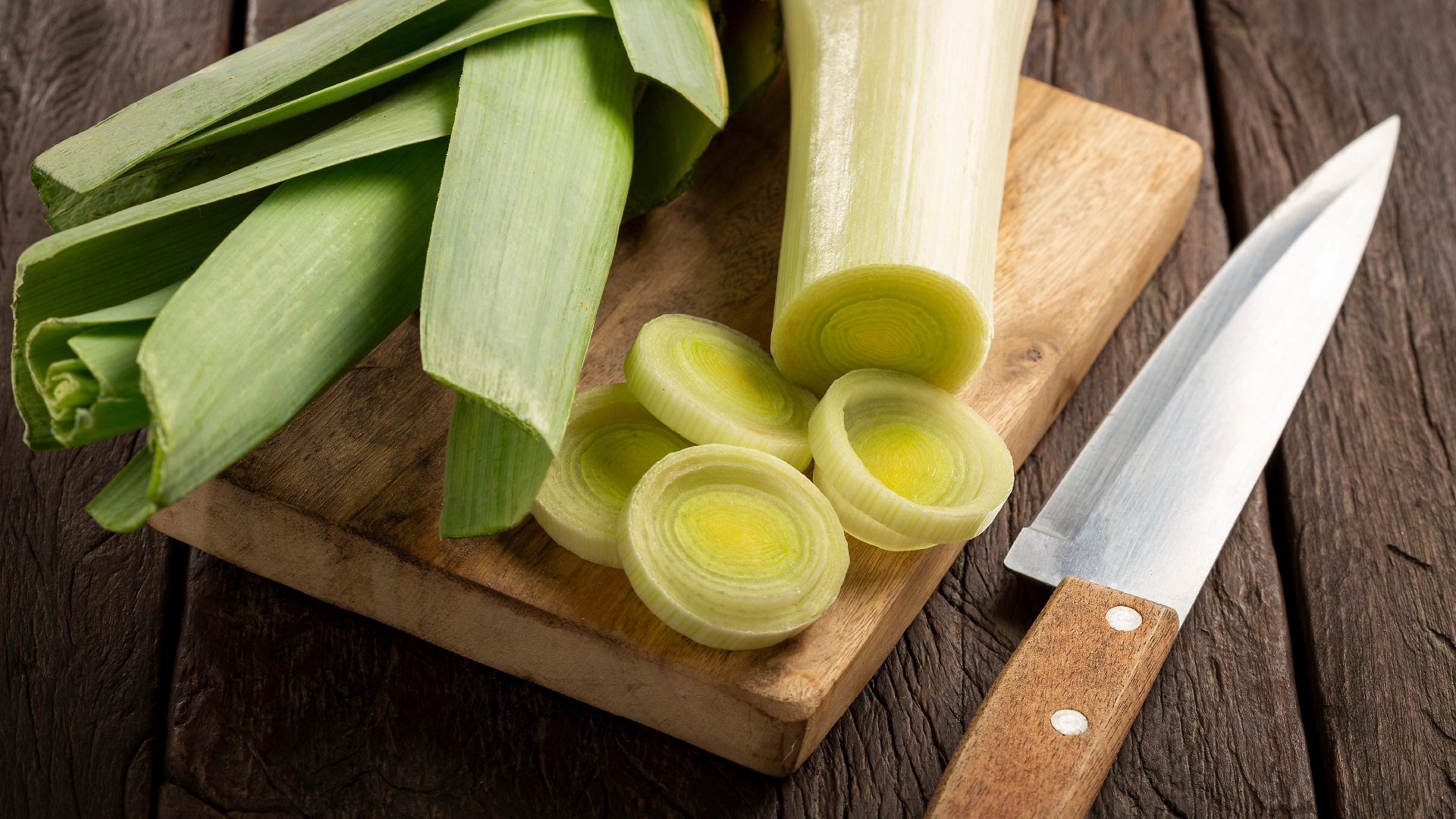 A leek sits next to four round slices on a wooden chopping board, with a sharp knife positioned to the right on the table. 