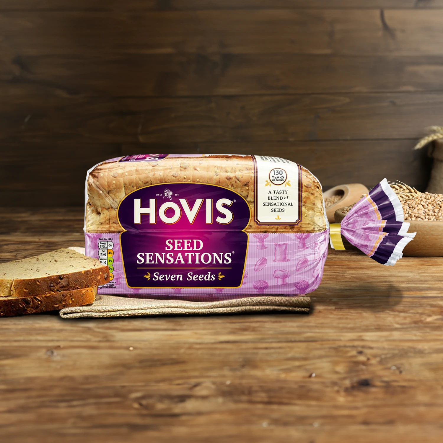Hovis Seed Sensations Original loaf on a wooden table, with a bowl of wheat grains, a wooden scoop, and dried wheat in the background, along with slices of bread on the left.