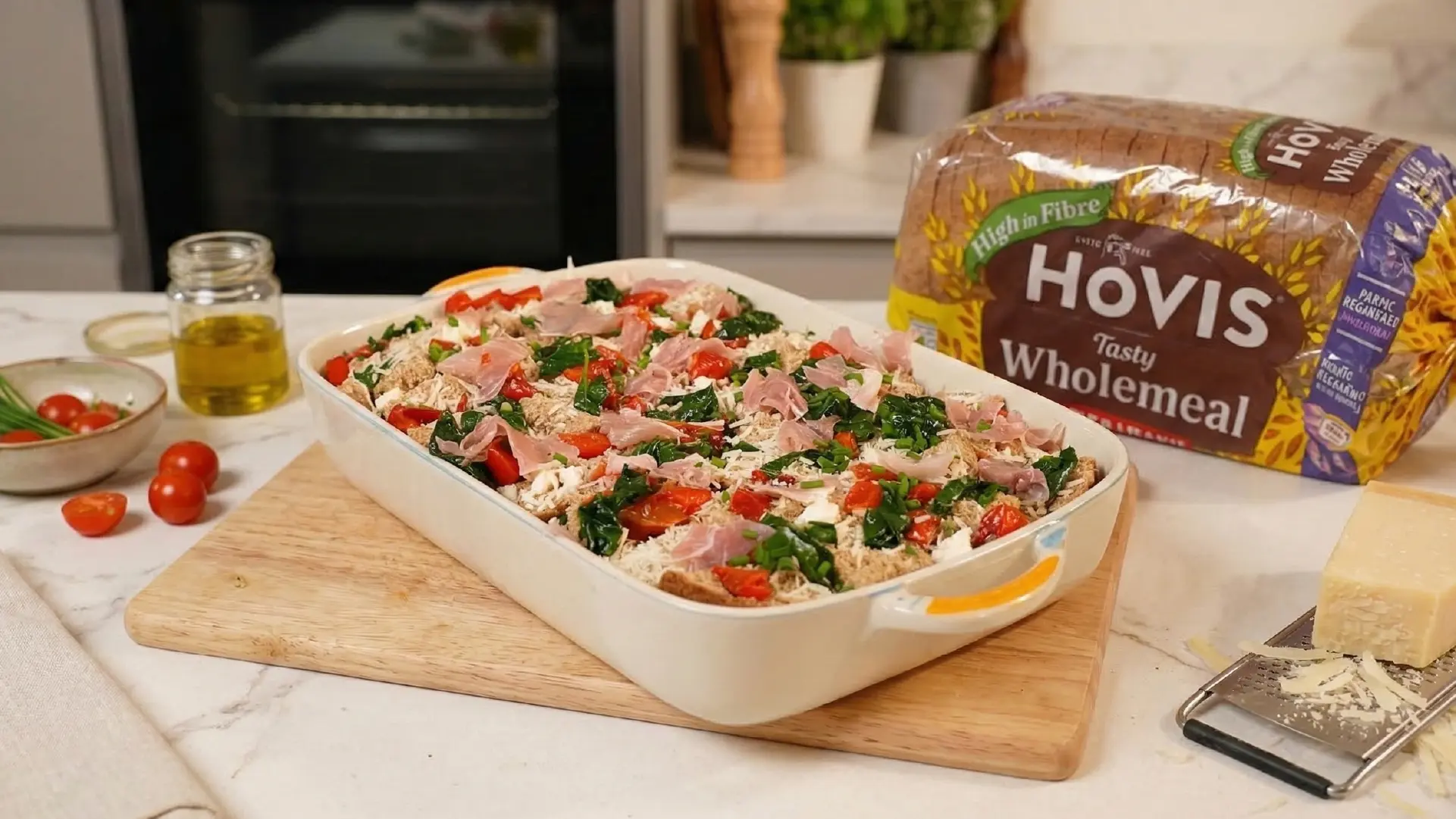 Unbaked breakfast strata layered with wholemeal bread, spinach, prosciutto, and tomatoes in a dish with cheese nearby.