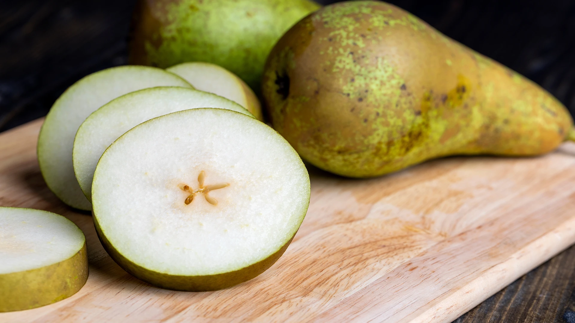 Five pear slices rest on a wooden chopping board. There are two whole green pears on the background, about to be sliced. 