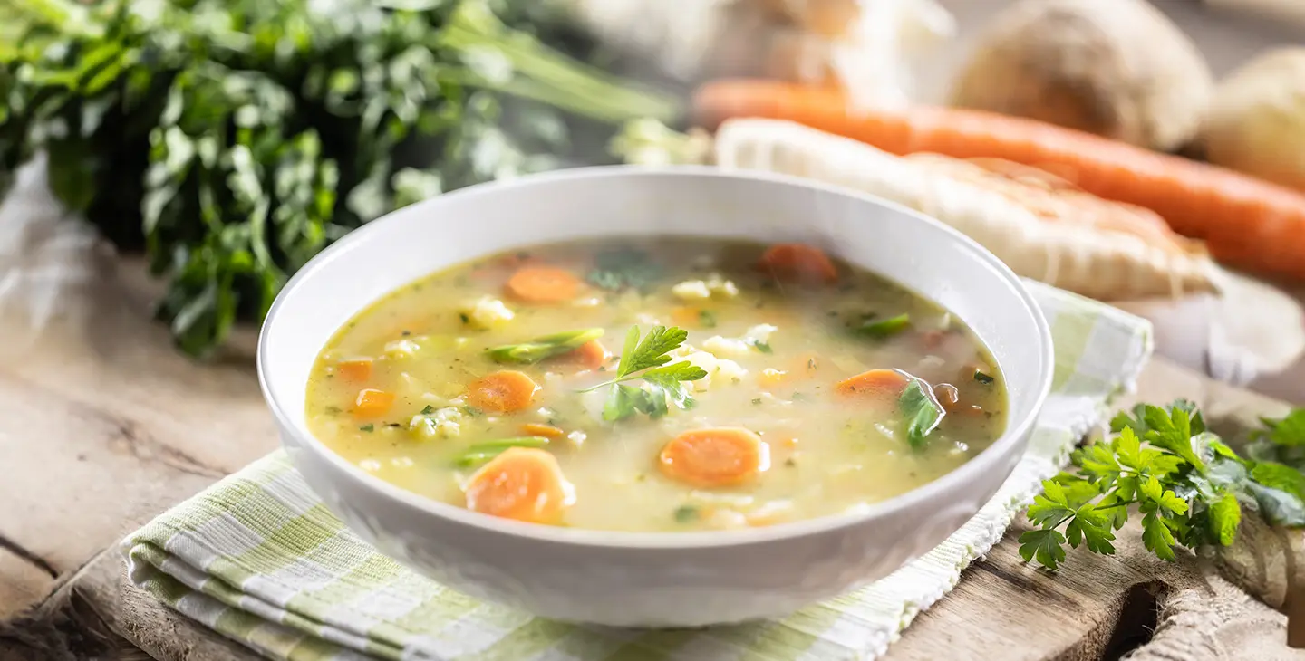 A steaming bowel of vegetable soup sits on a table cloth on a table surrounded by ingredients. 