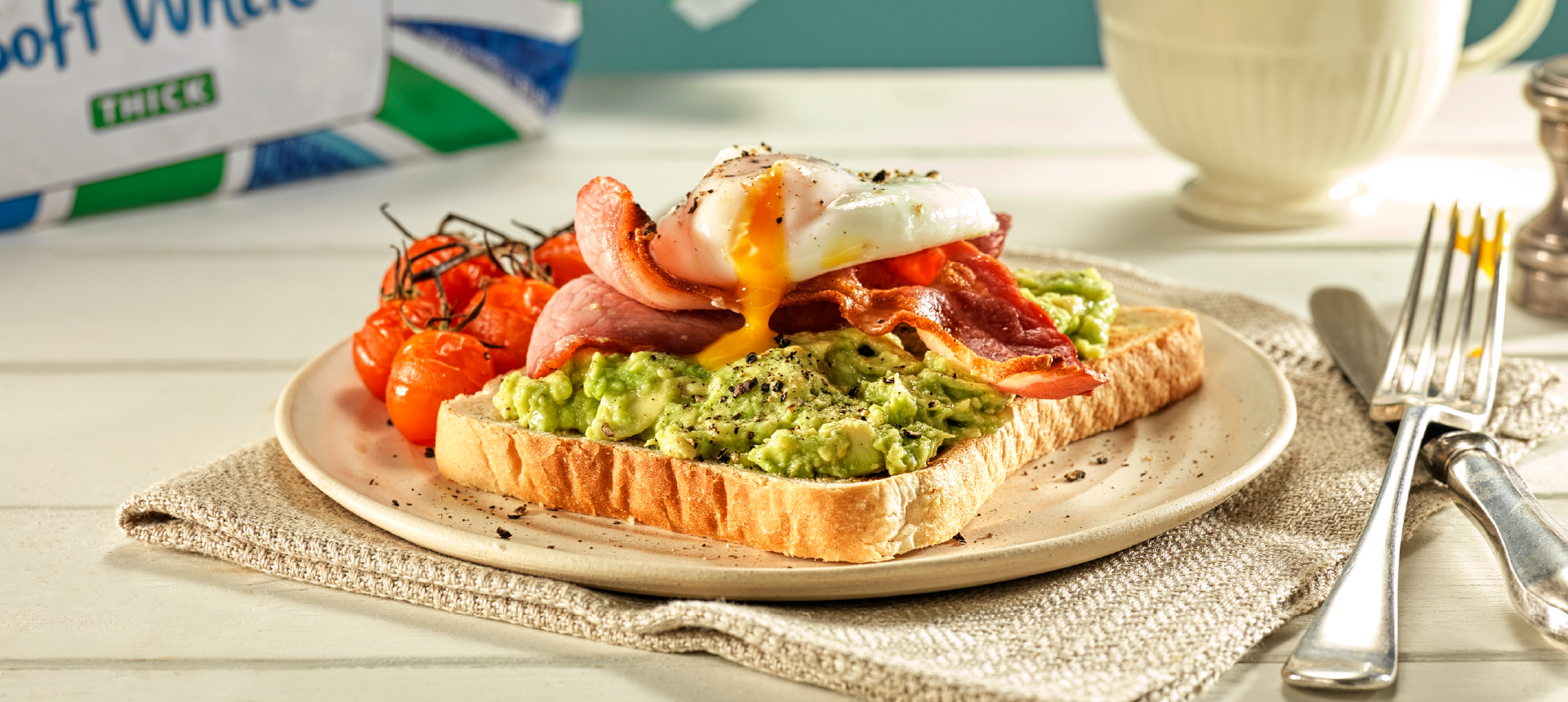 A slice of Hovis Soft White Thick Sliced on a plate, topped with mashed avocado, crispy bacon, a poached egg with yellow yolk running and some grilled vine cherry tomatoes on the side. 