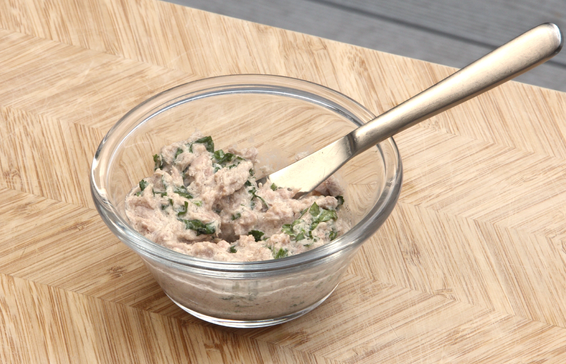 A small glass jar filled with a mix of tuna, mayo and chopped coriander, accompanied by a knife on top of a wooden chopping board.