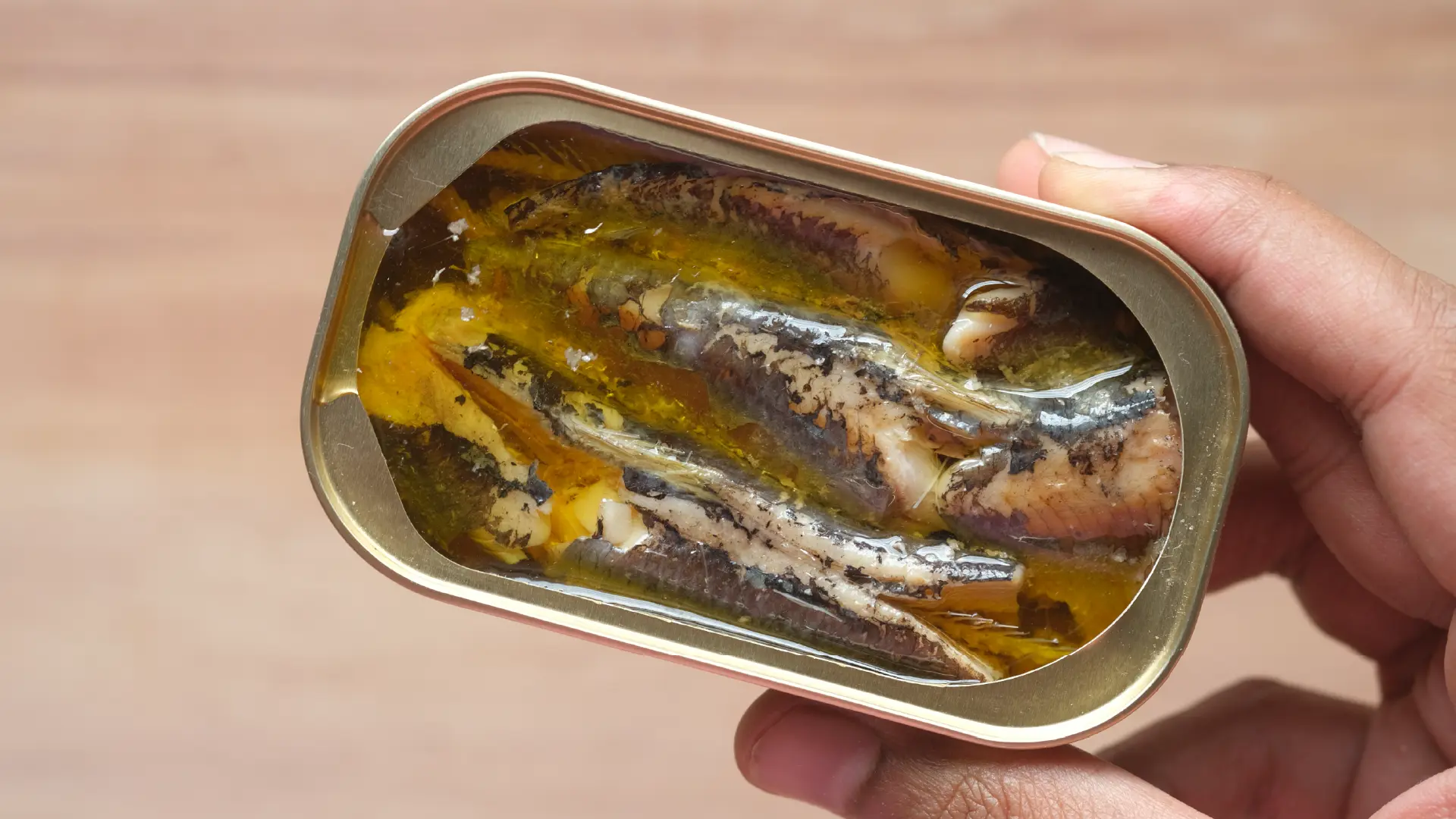 A hand holding an open rectangular tin of sardines packed closely together in olive oil, over a blurred wooden background.