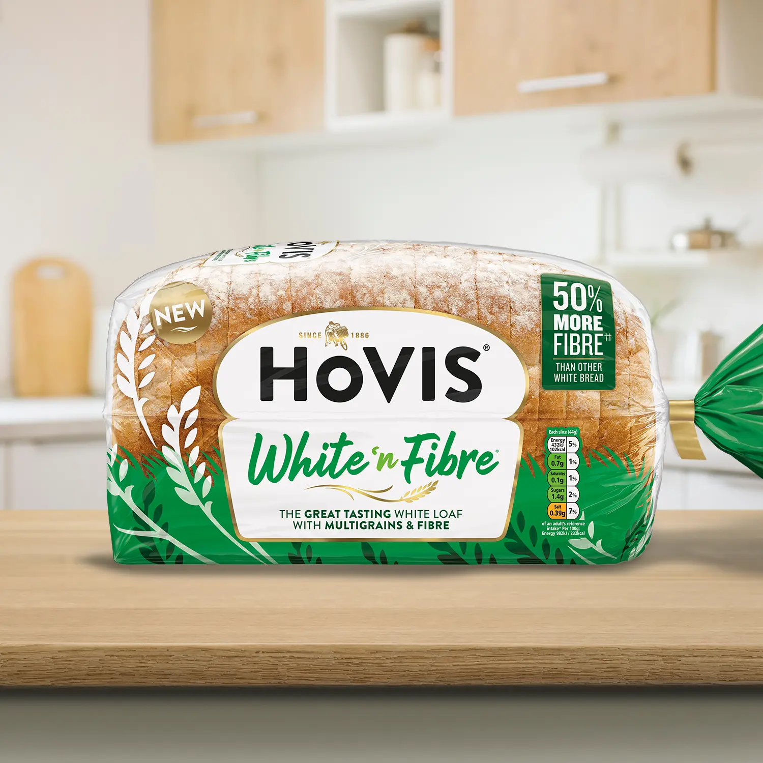 A loaf of Hovis White n Fibre bread with a crust dusted with flour, displayed on a wooden kitchen countertop. The packaging features a white and green design with a gold trim and nutrition information. A 'NEW' label is shown on the top corner of the pack
