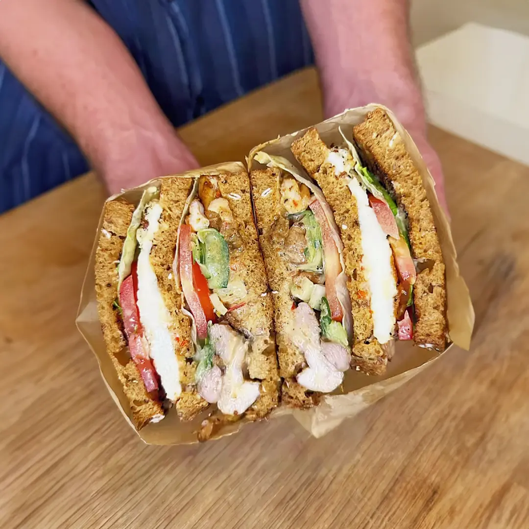 Someone holding two halves of a sandwich made with Hovis Granary Half Cob wrapped in paper. The sandwich is made of brown bread, filled with tomato slices, lettuce, juicy grilled chicken, and thin cucumber slices.