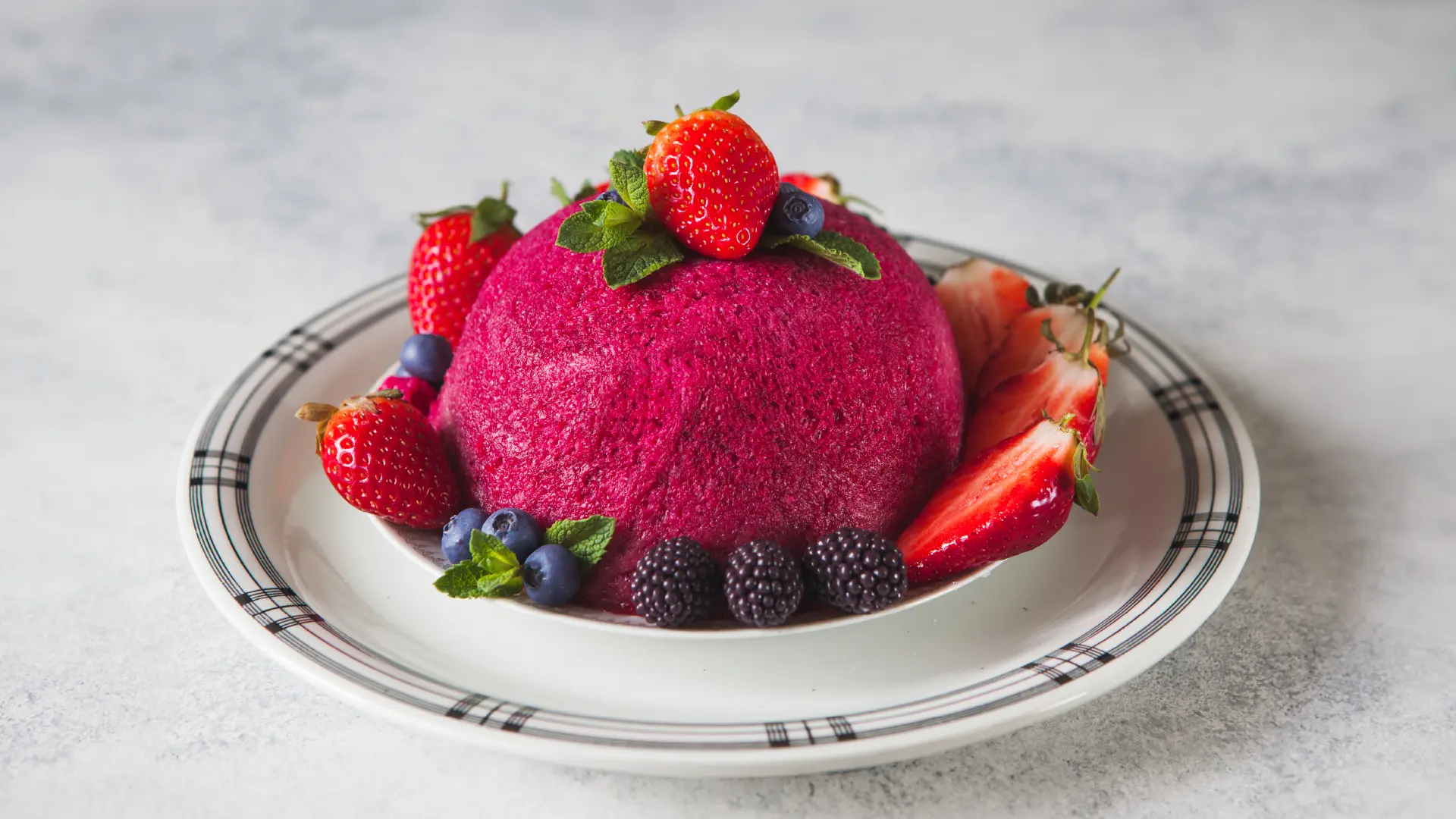 A small, bright pink dome-shaped bread pudding made with Hovis Farmhouse Soft White on a plate, topped with a strawberry and mint leaves. The base is surrounded by halved strawberries, blackberries, blueberries, and more mint leaves.