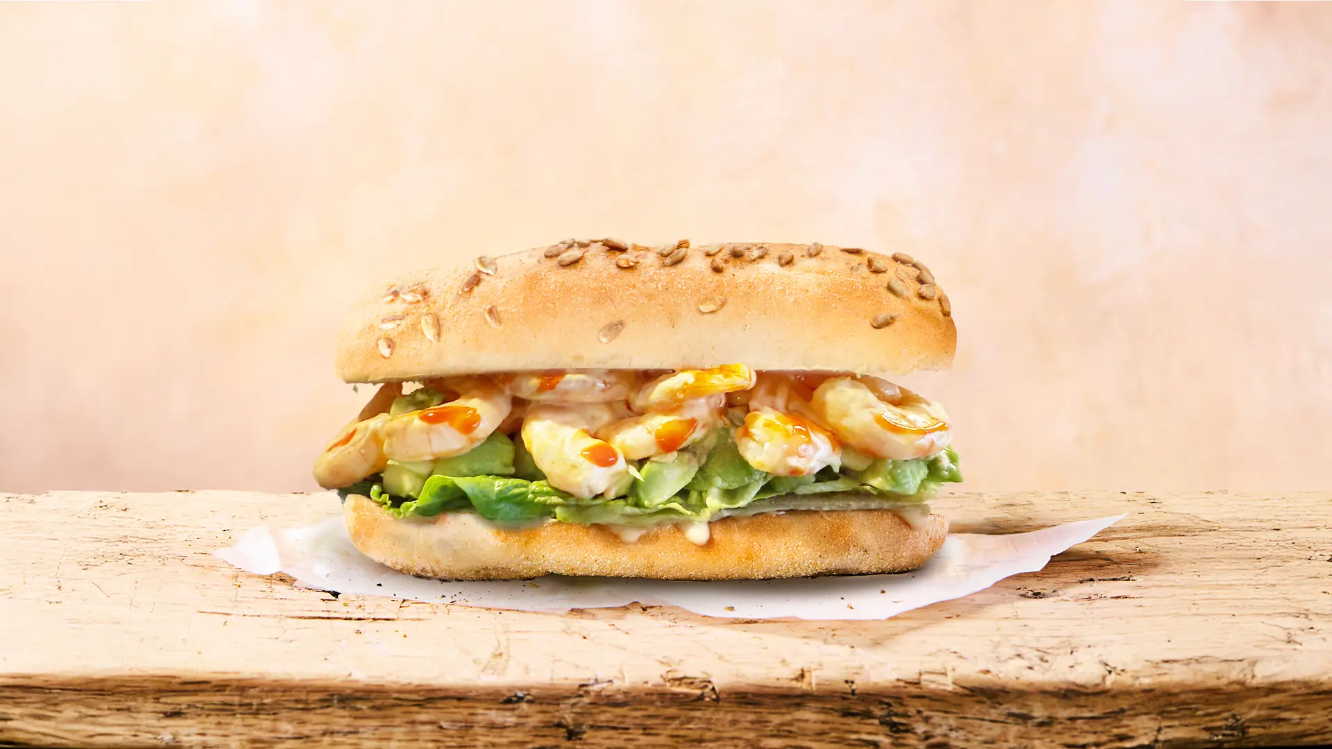 A fresh Protein Sunflower & Honey Sub Rolls sandwich, filled with medium sized prawns, green leaves, melded cheese and chilli sauce rest on top of a napkin in a light beige background. 