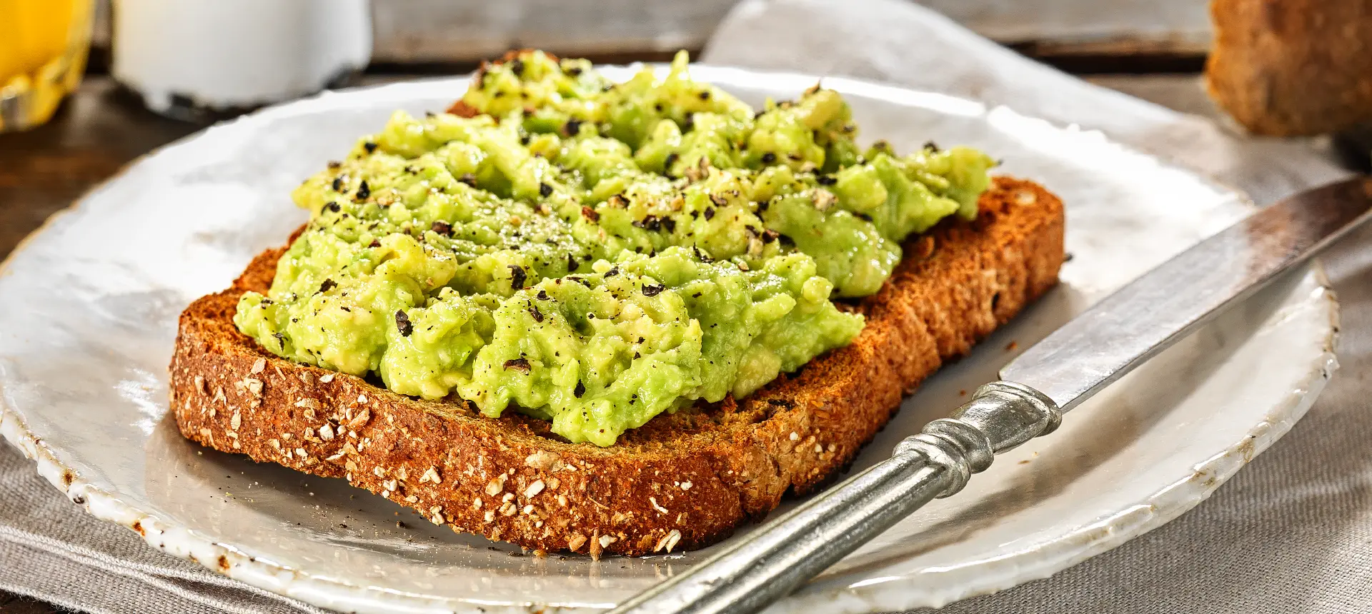 A slice of Hovis Granary Wholemeal bread topped with smashed avocado and a sprinkle of ground black pepper, on a plate with a butter knife on the side.
