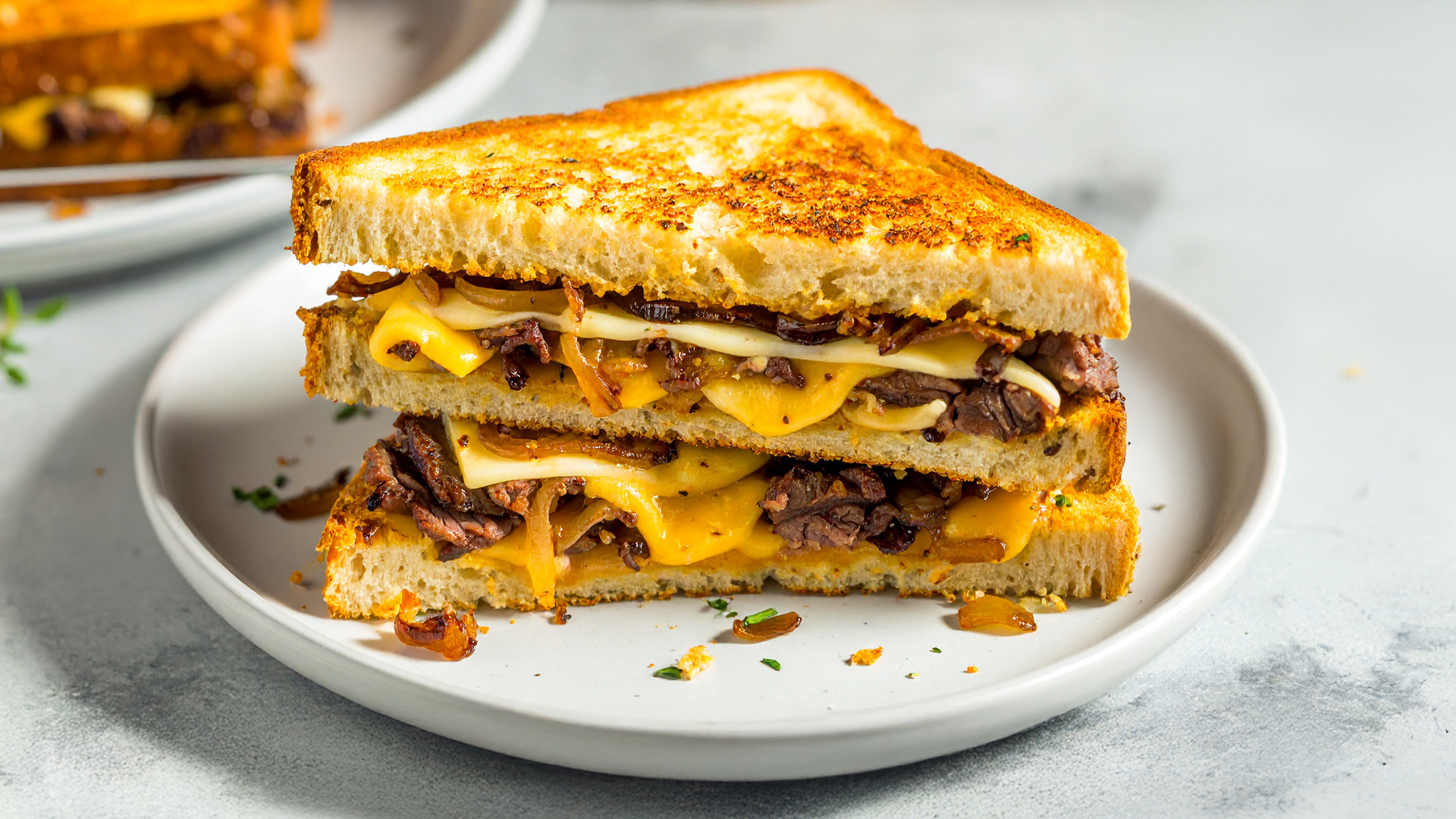 A delicious Beef & Caramelised Onion Grilled Cheese sandwich cut in half on a white plate, on a marbled table. In the background, another sandwich on a plate. 