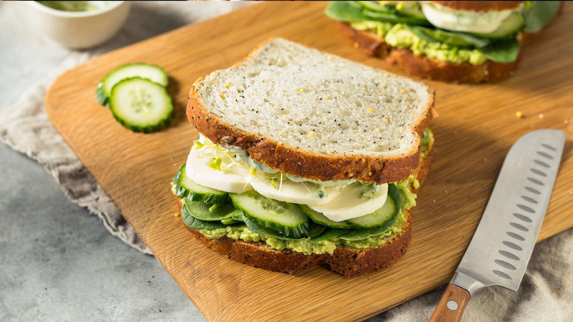 A Hovis Seed Sensations Original sandwich filled with mashed avocado, sliced cucumber, and mozzarella cheese, with cucumber slices and a knife on the side.