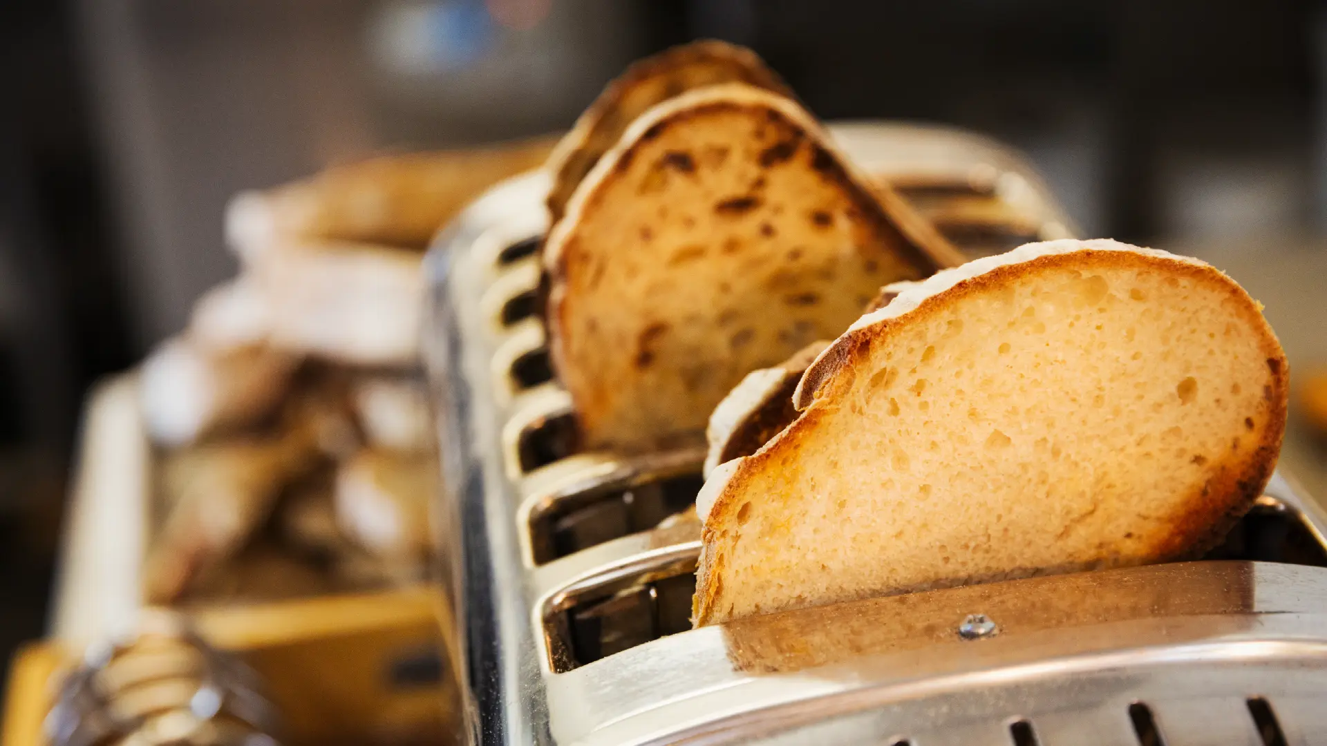 Slices of rustic sourdough bread being toasted in a commercial, multi-slot stainless steel toaster in a bakery.