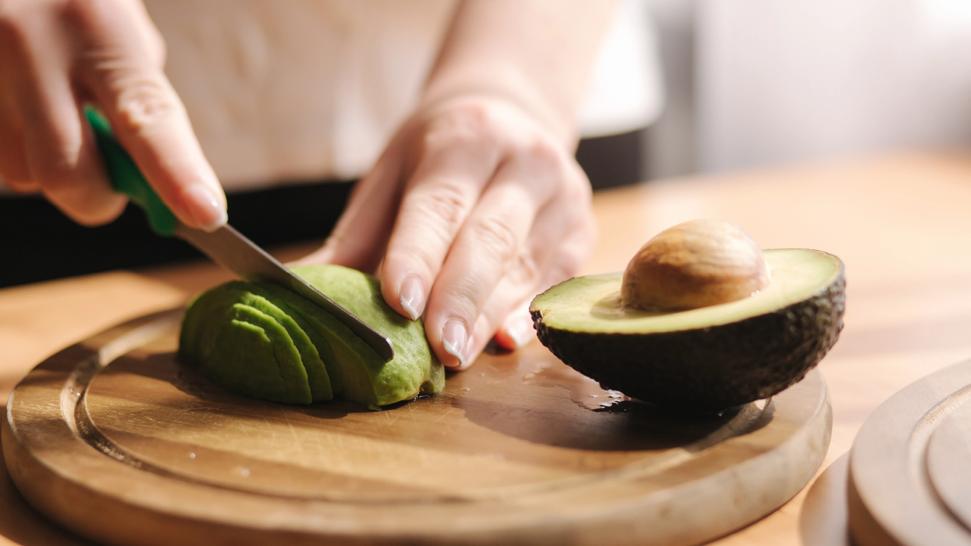 Hands slicing one half of an avocado lengthwise, with a small knife, on a wooden chopping board, with the seeded half resting on the right.