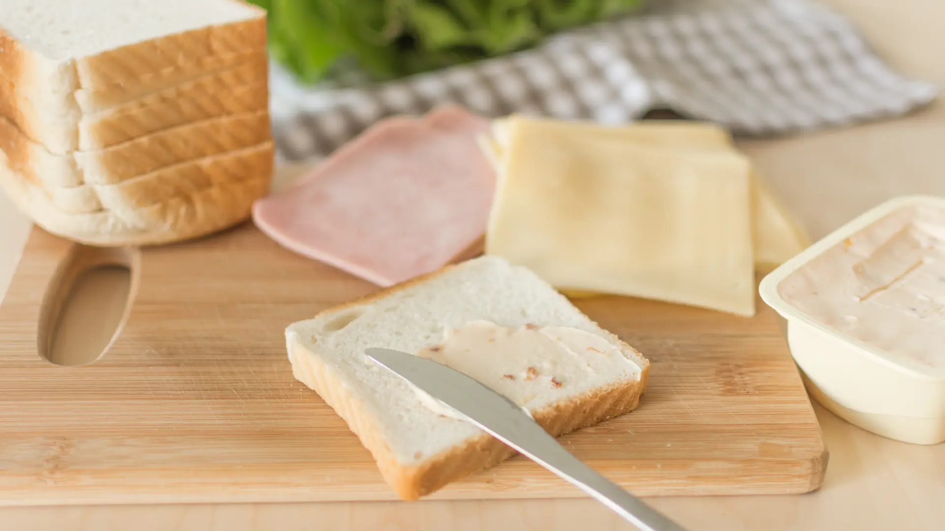 A knife spreading butter on a slice of Hovis Soft White Thick Sliced bread in the kitchen on a wooden chopping board surrounded by more bread and other ingredients.