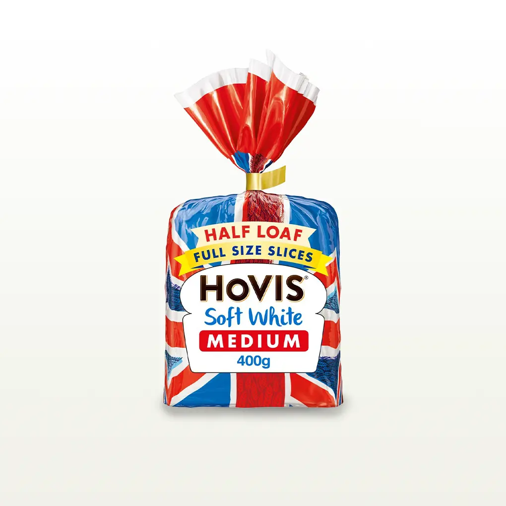 Hovis Soft White Medium Sliced 400g bread on a white backdrop with a subtle shadow. The packaging features a Union Jack pattern and a yellow label reading 'Full Size Slices.'