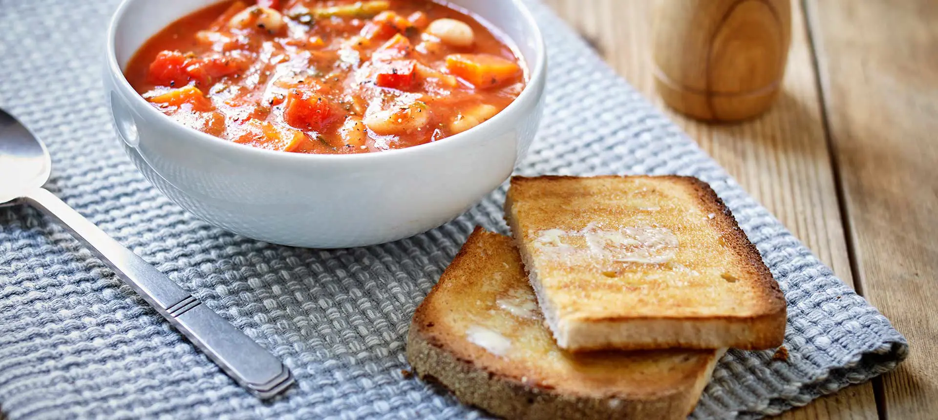 Two toasted slices of buttered Mothers Pride Scottish Plain bread on a blue tablecloth, with a bowl of vegetable soup and a spoon on the side.