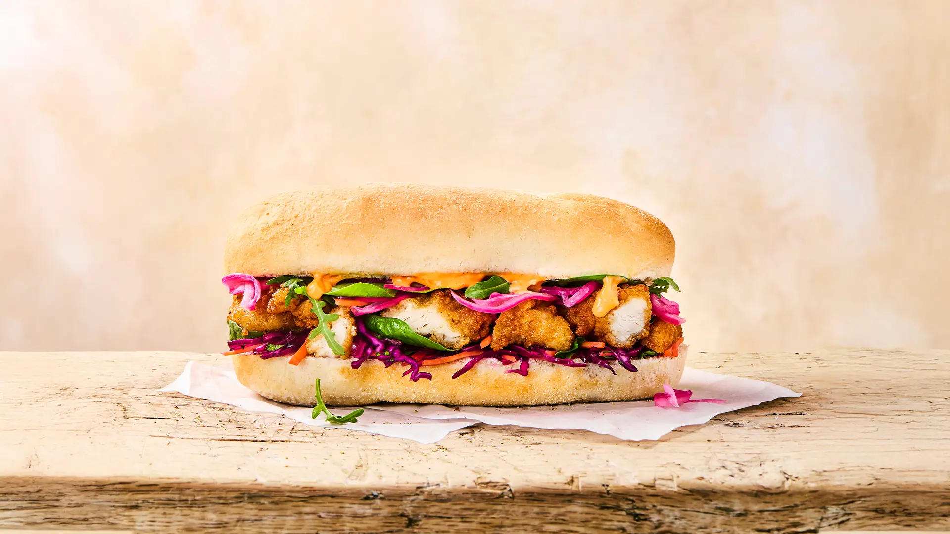 A fresh Hovis Buttermilk Sub Rolls sandwich, filled with pieces of fried chicken, grated red cabbage and carrots, rockets and sauce rest on top of a napkin in a light beige background. 