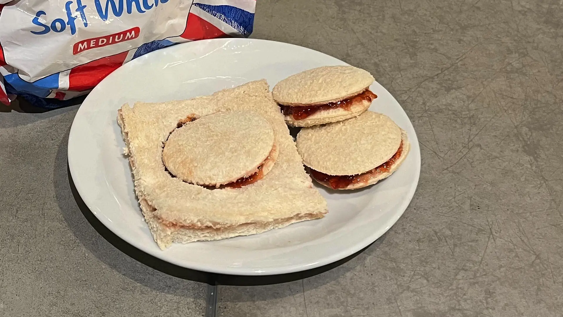 Two small, circular jam sandwiches made with Hovis Soft White Medium Sliced bread sit on a plate, along with a half-cut jam penny still in its square slices of bread, yet to be unmoulded.