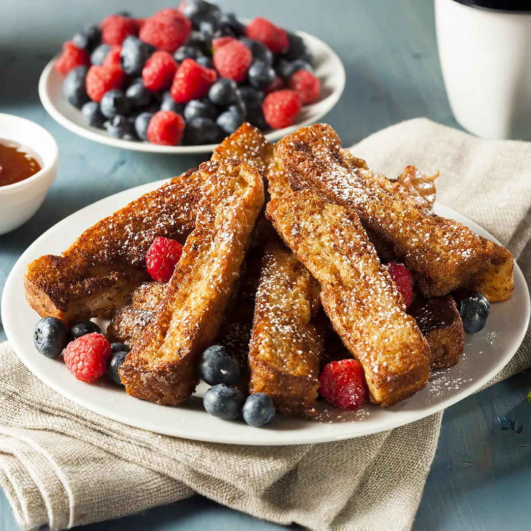  Golden brown bread sticks topped with sugar, surrounded by berries on a white plate. In the background some extra berries and a small bowl with chocolate dip. 