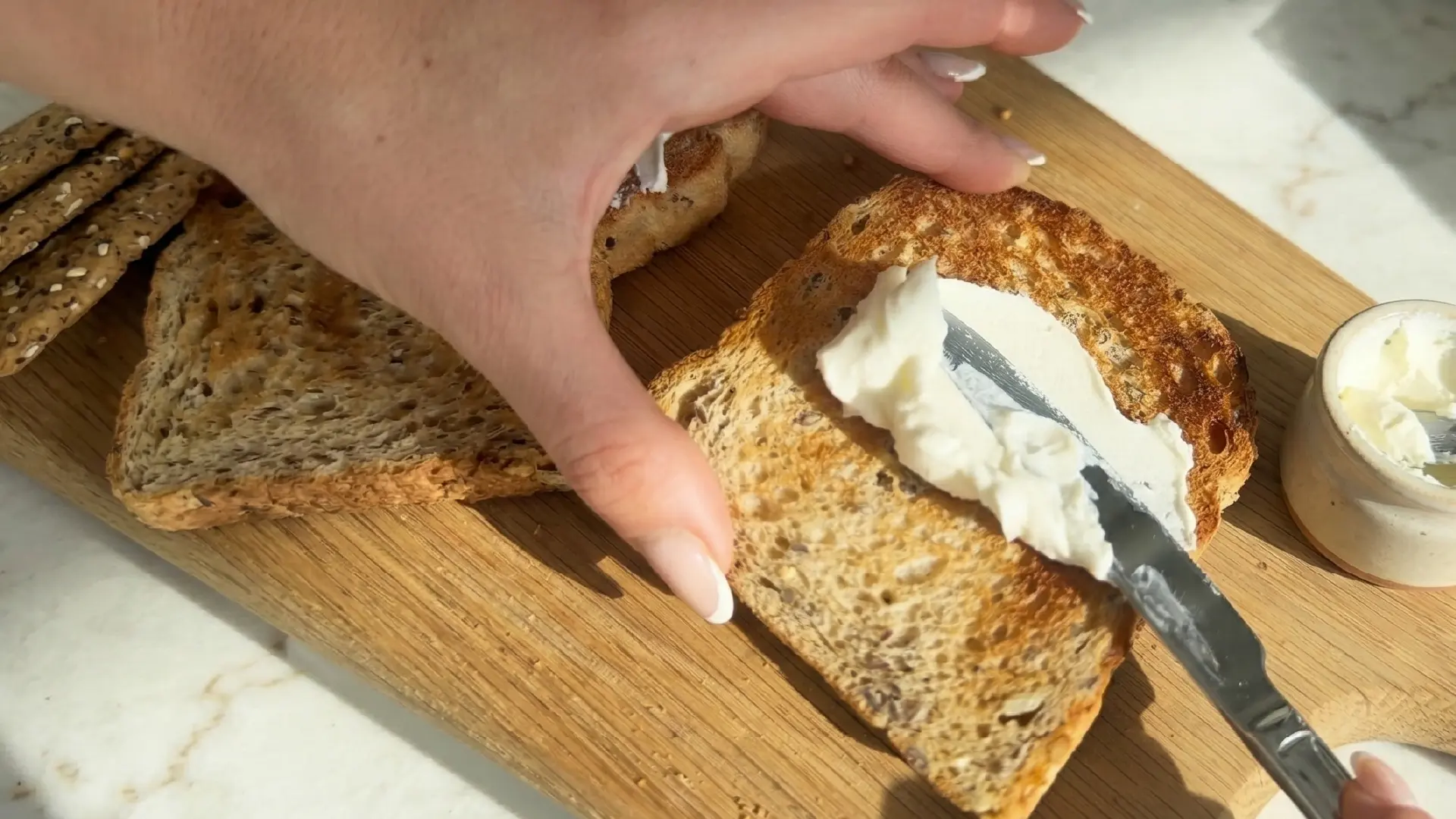 A close-up of a hand using a butter knife to spread thick white cream cheese onto a slice of golden-brown seeded toast.